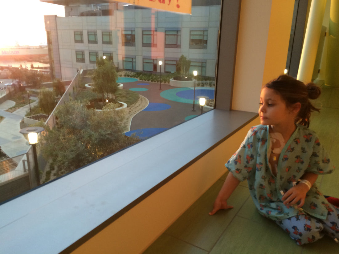 Picture of a little girl in hospital pajamas with tubes taped to her chest, looking out the window as the sun rises