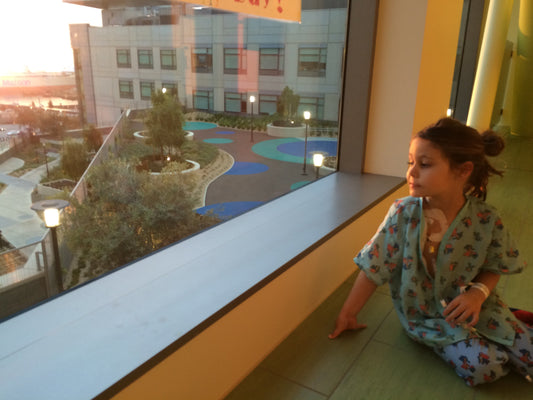 Picture of a little girl in hospital pajamas with tubes taped to her chest, looking out the window as the sun rises