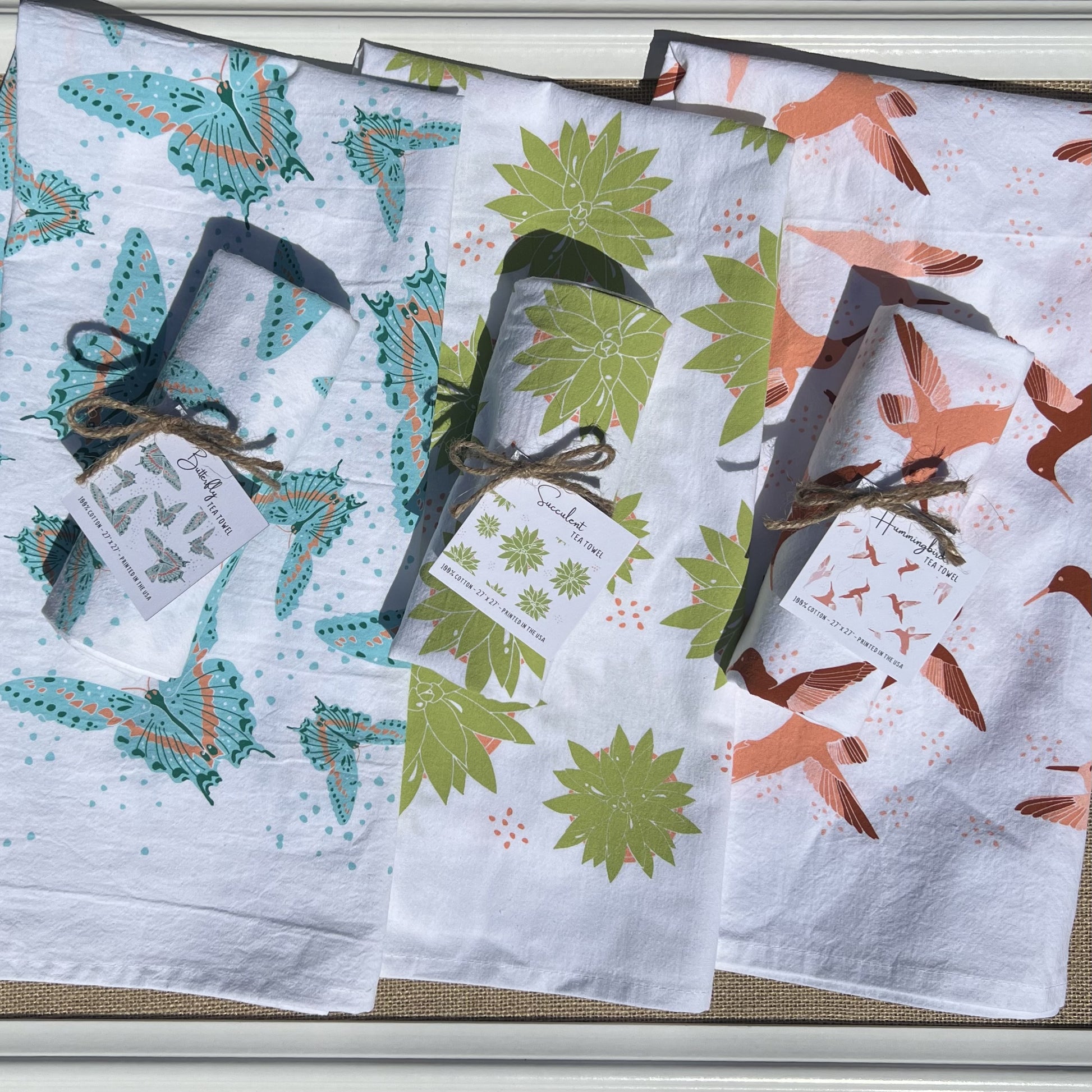 Line up of three tea towel prints, all on white backgrounds: blue butterfly, green succulent and pink hummingbird. Shown with one towel of each color rolled up and one laid out to showcase the print.