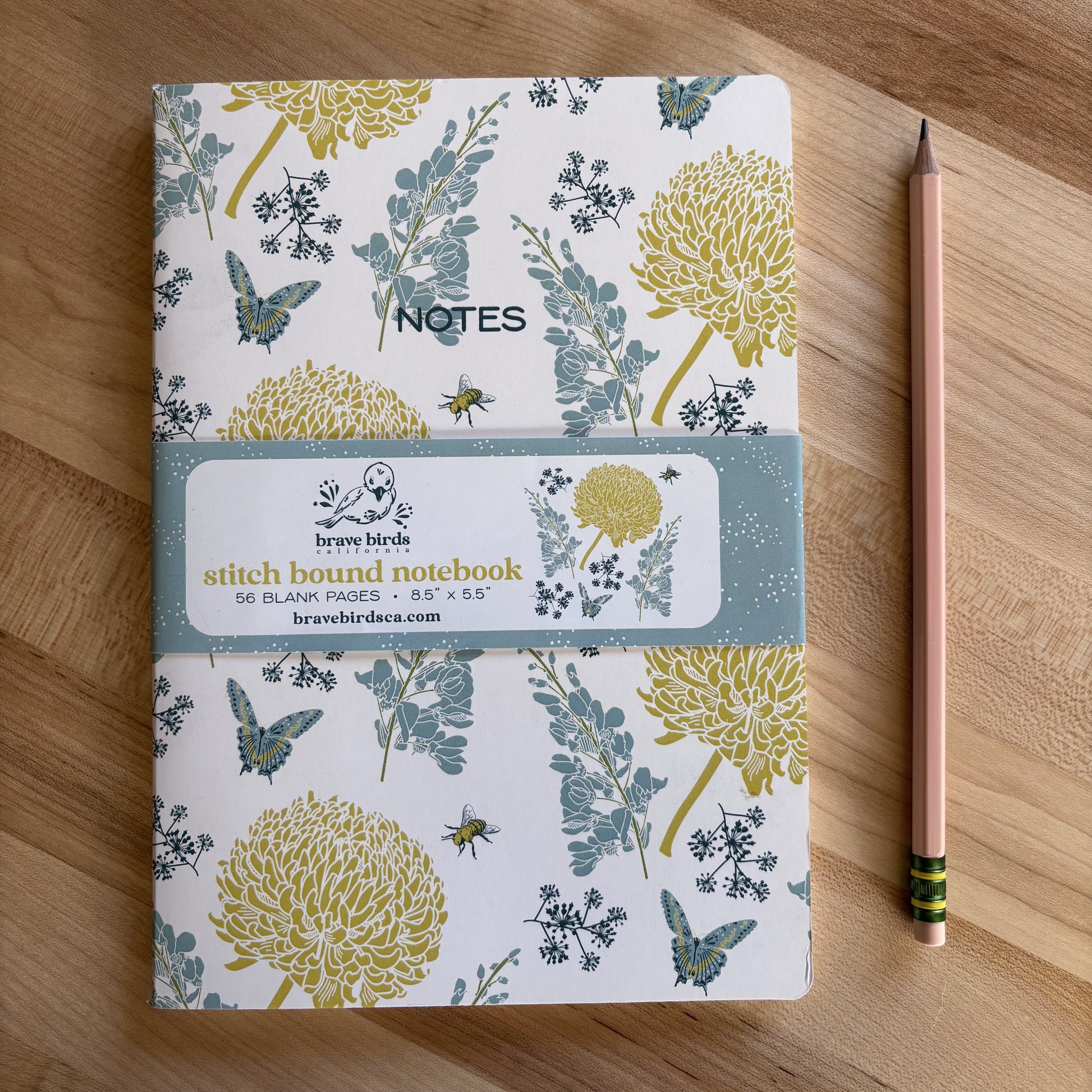 Blue and yellow floral print notebook with paper branded belly band, shown on a wood table with a pencil