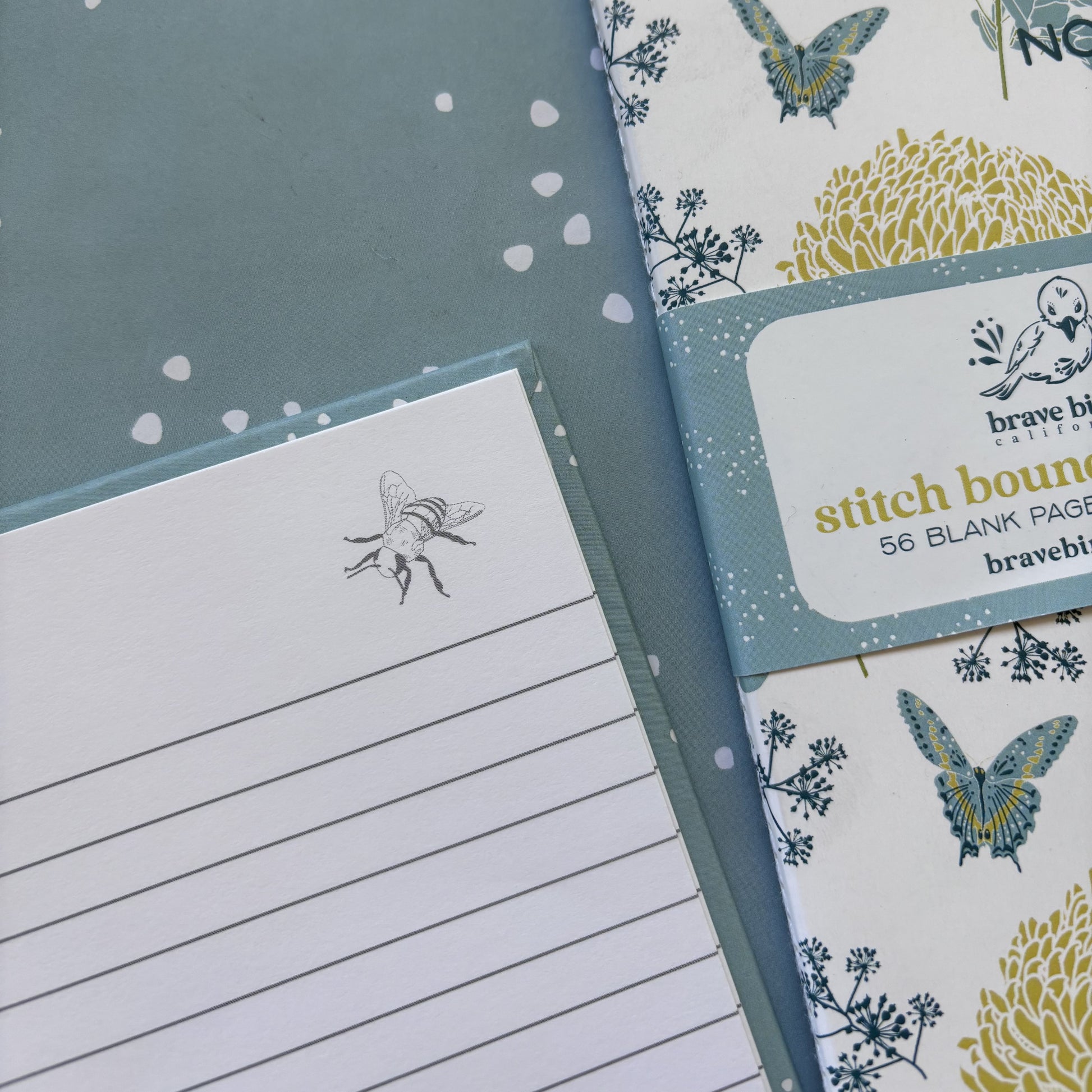 Close up of notebook with lines and bee in the corner & blue and yellow floral print notebook with paper branded belly band, shown on a blue dot background