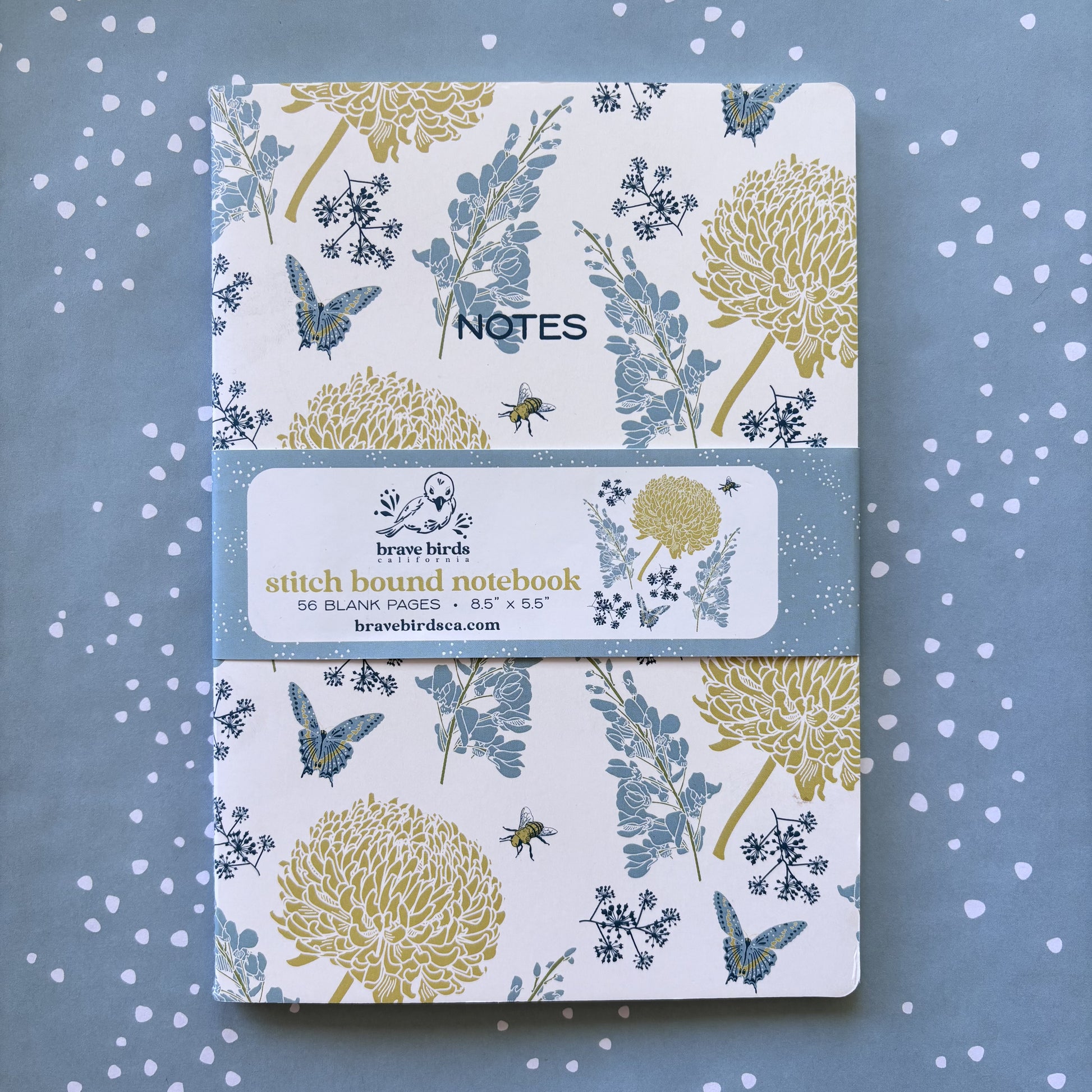 Blue and yellow floral print notebook with paper branded belly band, shown on blue and white dot background.