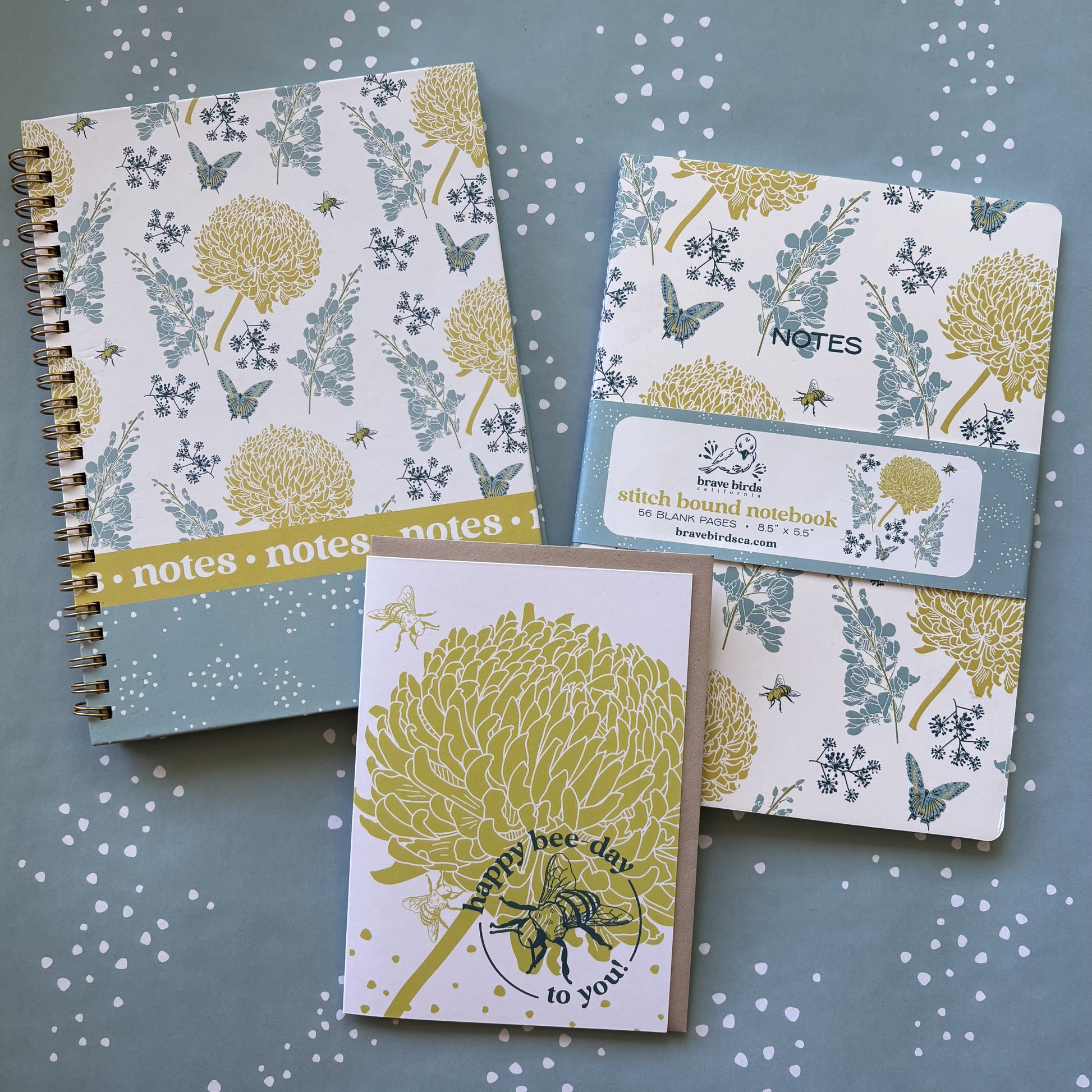 Blue and yellow floral print spiral notebook, blue and yellow floral print notebook with paper branded belly band, and card with yellow flower and bee shown on a blue and white dot background.
