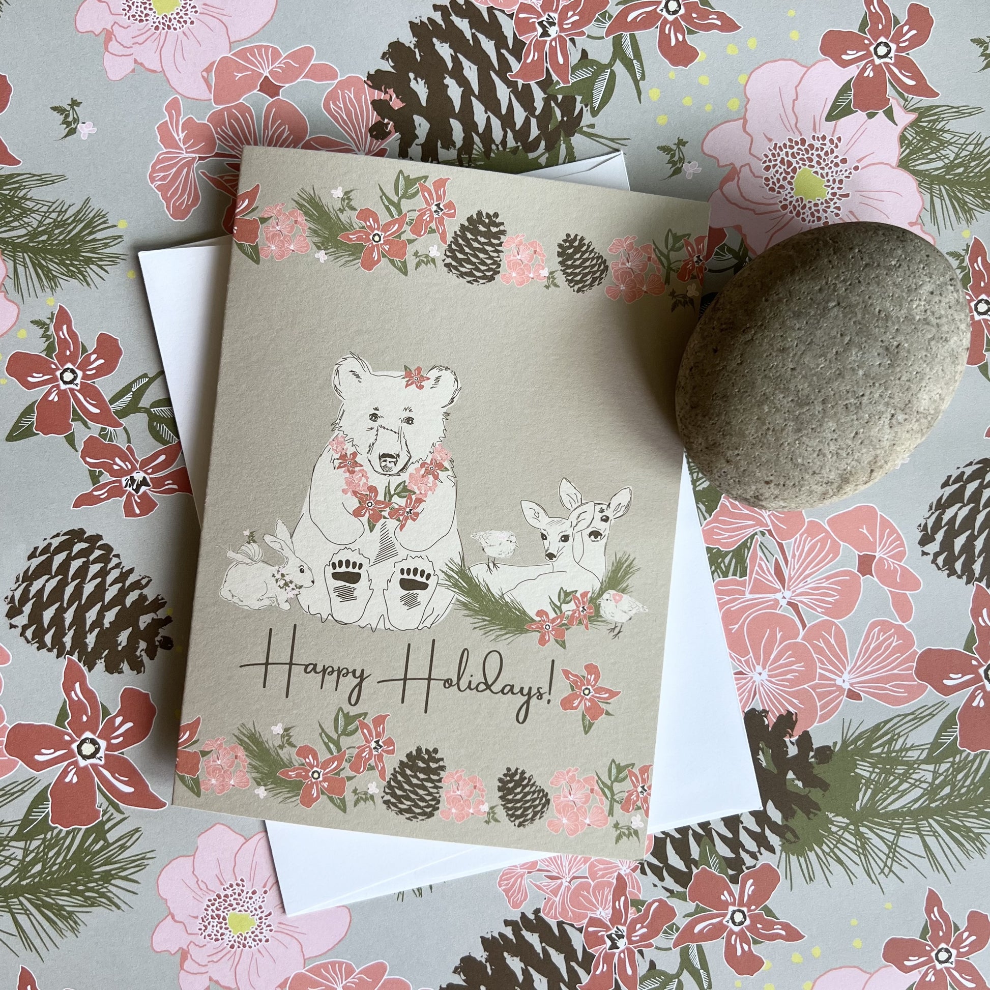 Holiday card with a kraft paper brown background and illustrations of a bear, deer, bunny, birds and a snail, all decorated with festive flowers. Text reads "Happy Holidays"