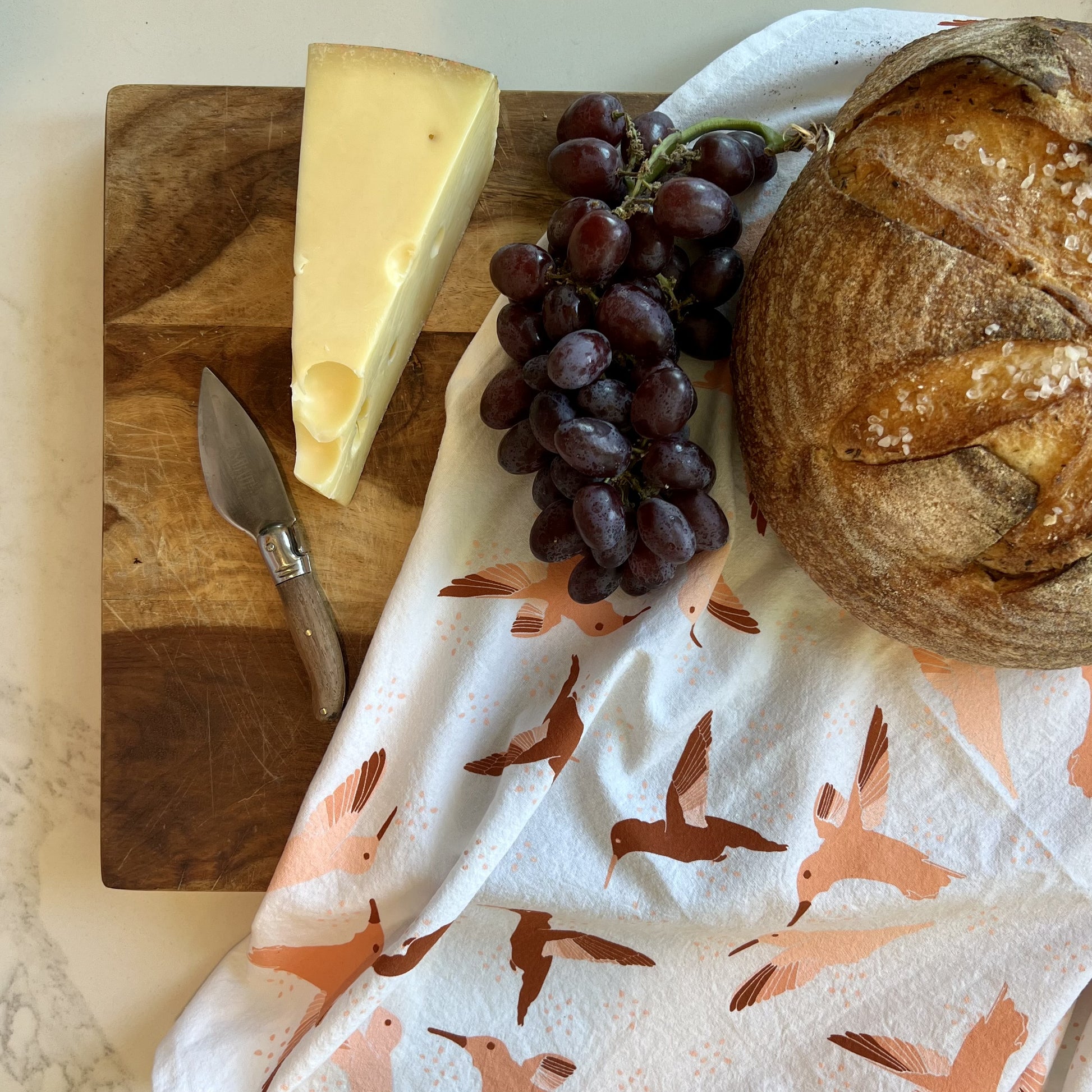 Hummingbird tea towel shown with a wood platter, bread, cheese and grapes