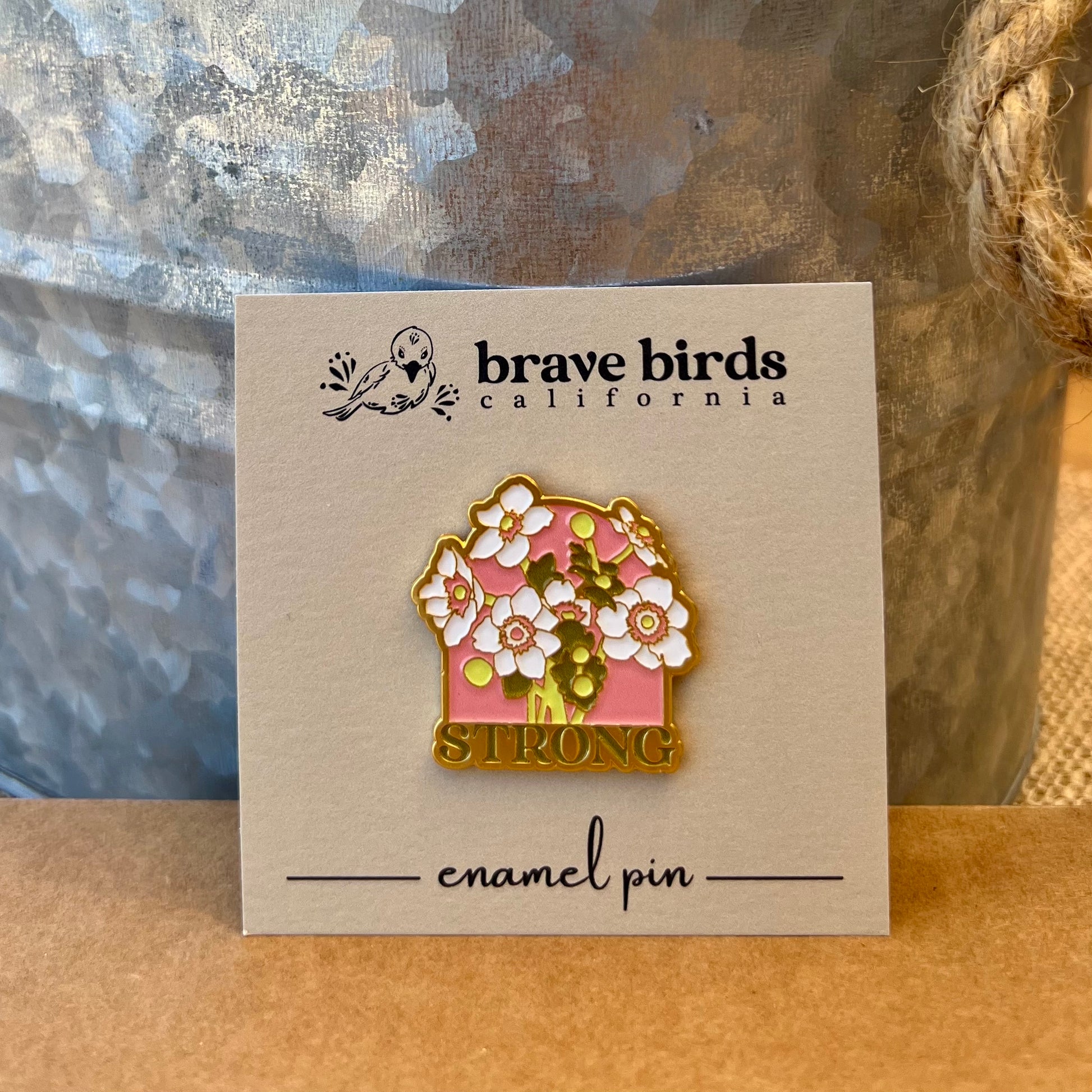 Enamel pin on a square backing card, featuring white flowers with pink & green accents and the word "STRONG"