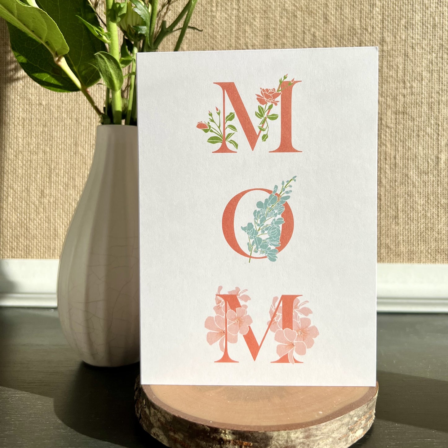 Greeting card with capital letters M-O-M in dark pink and flower illustrations wrapping around the letters, set on a desk with a wood coaster and a vase