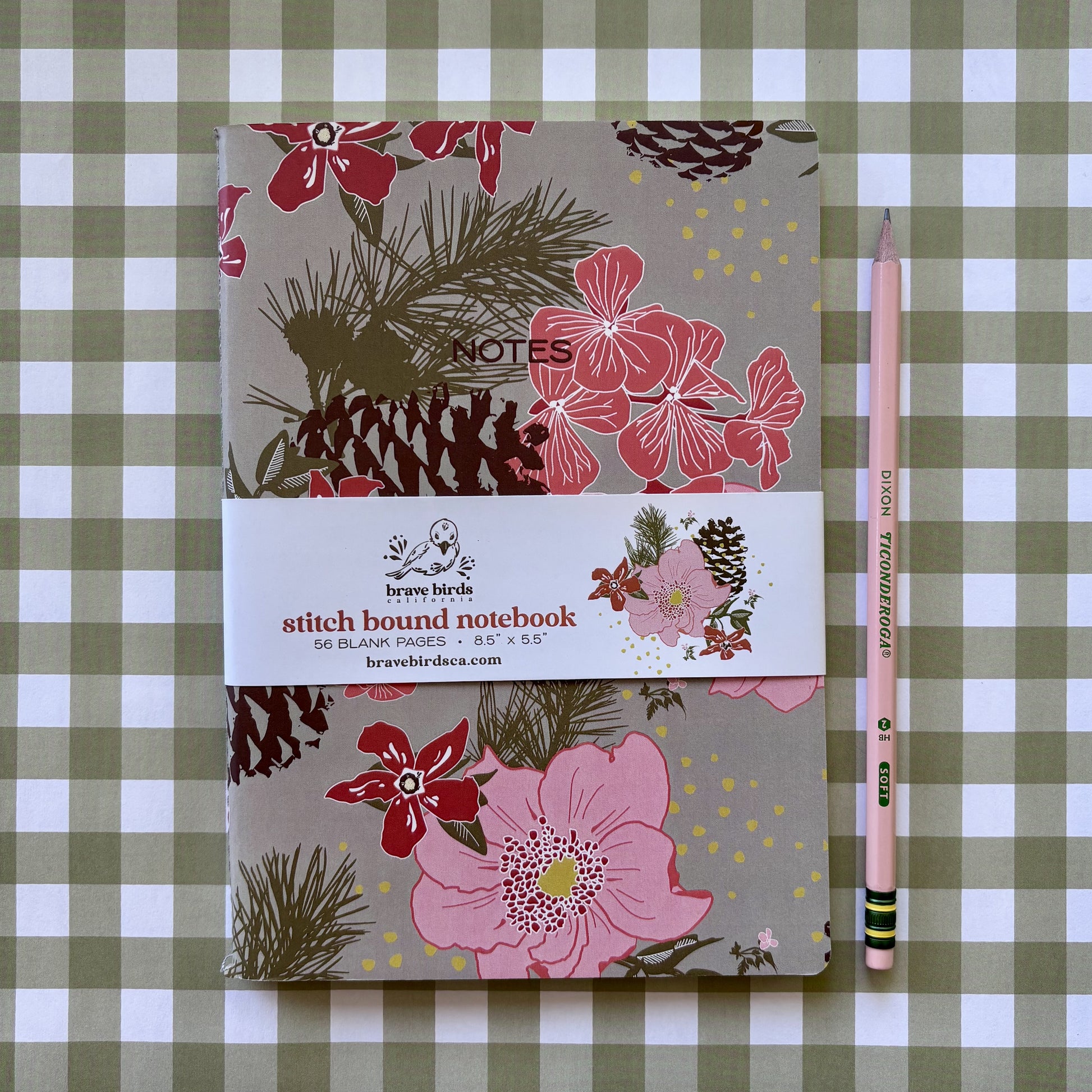 Pink, red and green floral print notebook with pinecones and paper branded belly band, shown on a green and white plaid background with a pencil