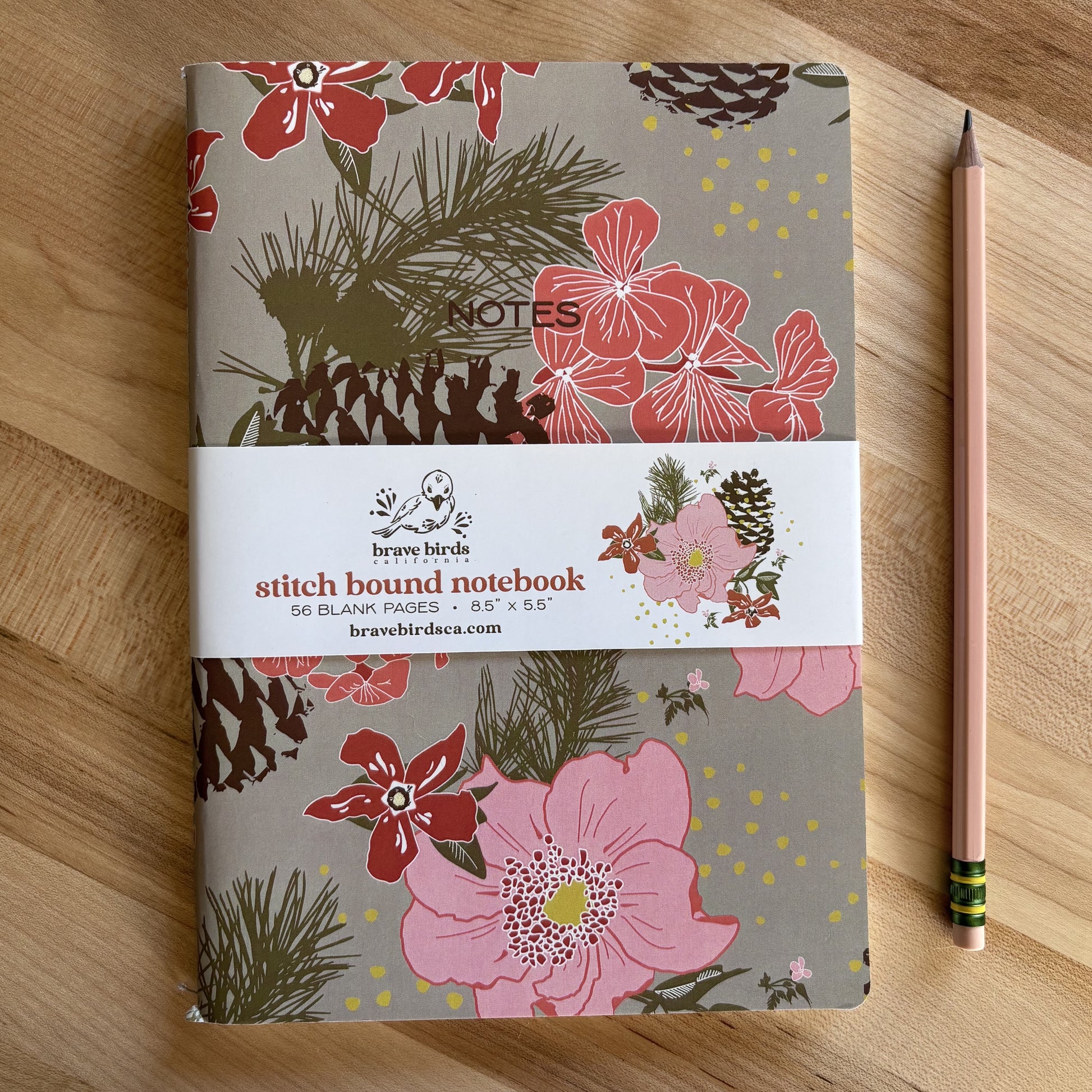 Pink, red and green floral print notebook with pinecones and paper branded belly band, shown on a wood table with a pencil