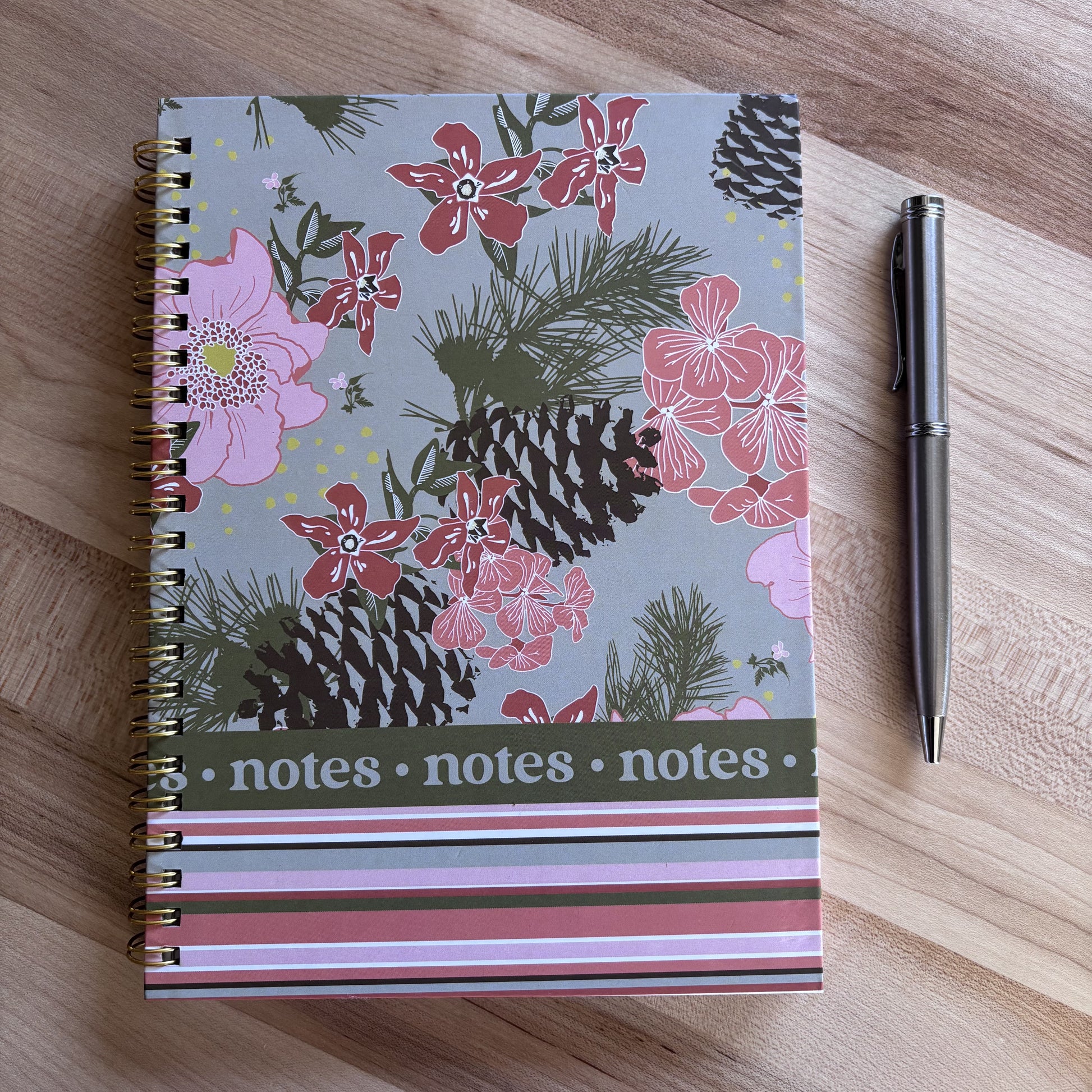 pink and green floral and stripe spiral notebook with pinecones on brown table with a pen