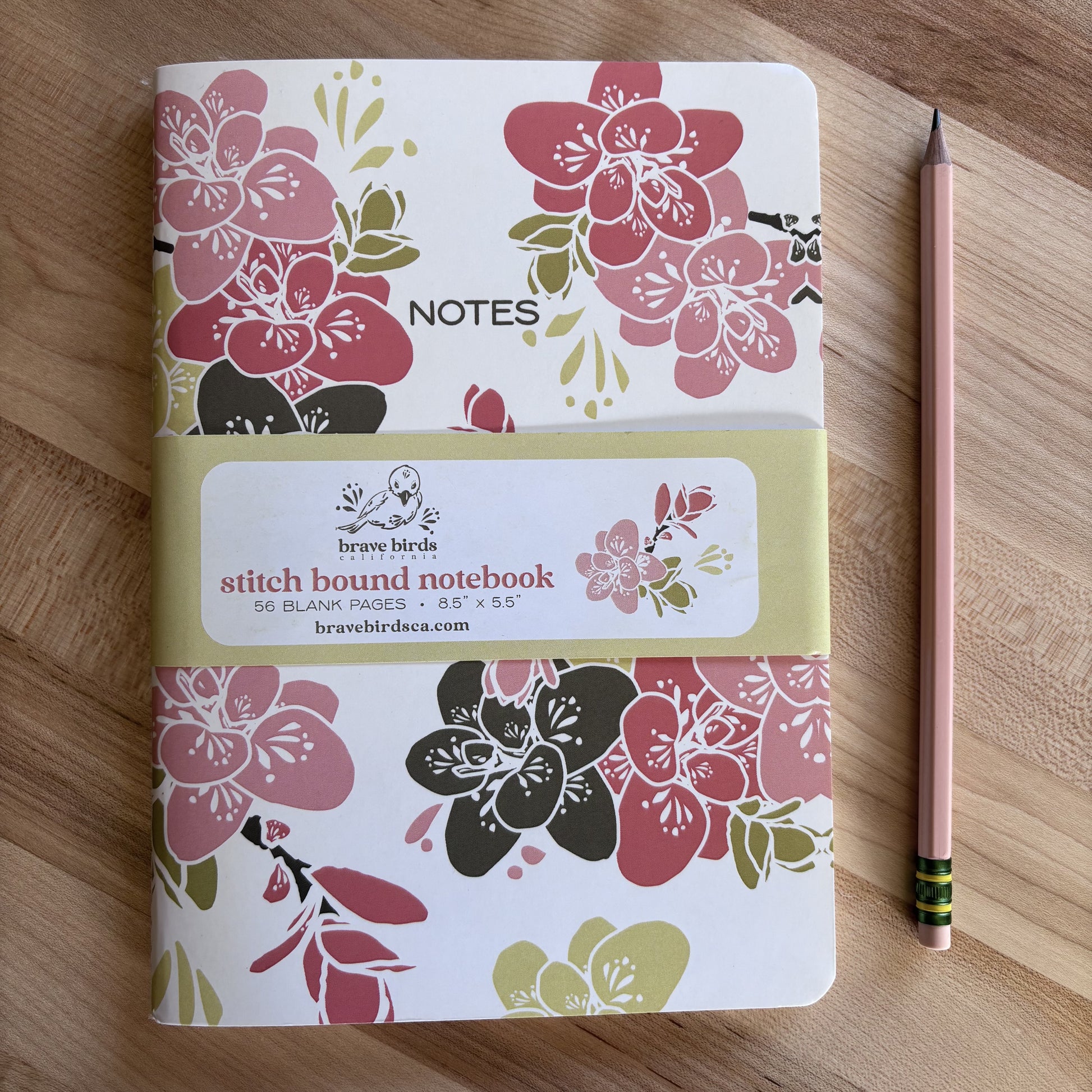 Pink and green floral print notebook with paper branded belly band, shown on a wood table with a pencil