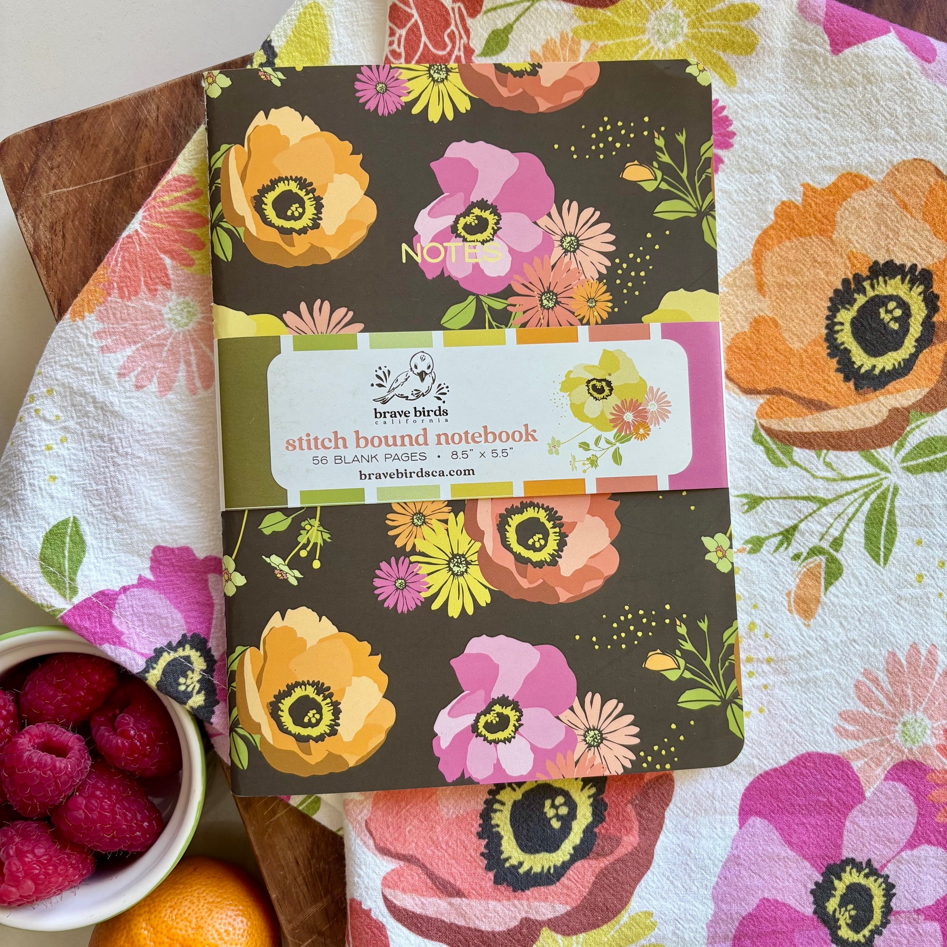 Floral notebook on a floral tablecloth with raspberries and an orange.