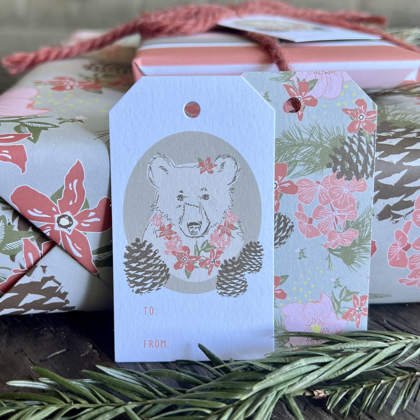 Gift wrapped package and two TO/FROM gift tags with an illustration of a bear with flower necklace and a flower in her ear on the front, an all-over floral print on the back..