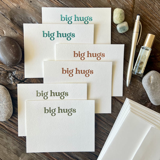 Staggered pile of six flat notecards, white in color with the words "big hugs" letterpress printed with embossed texture, two cards in teal ink, two in a warm orange and two in olive green. Styled on a wooden table with river rocks, a gold pen and a bottle of essential oil.
