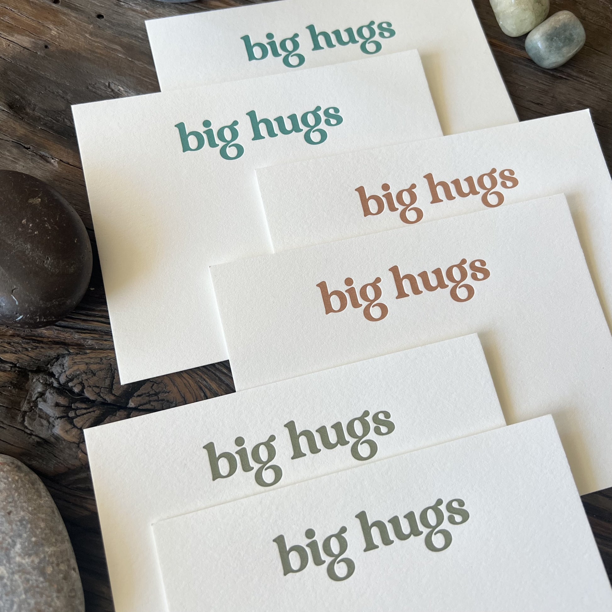 Closeup shot showing debossed texture of letterpress printed notecards that say "big hugs" in teal, olive and dark orange.