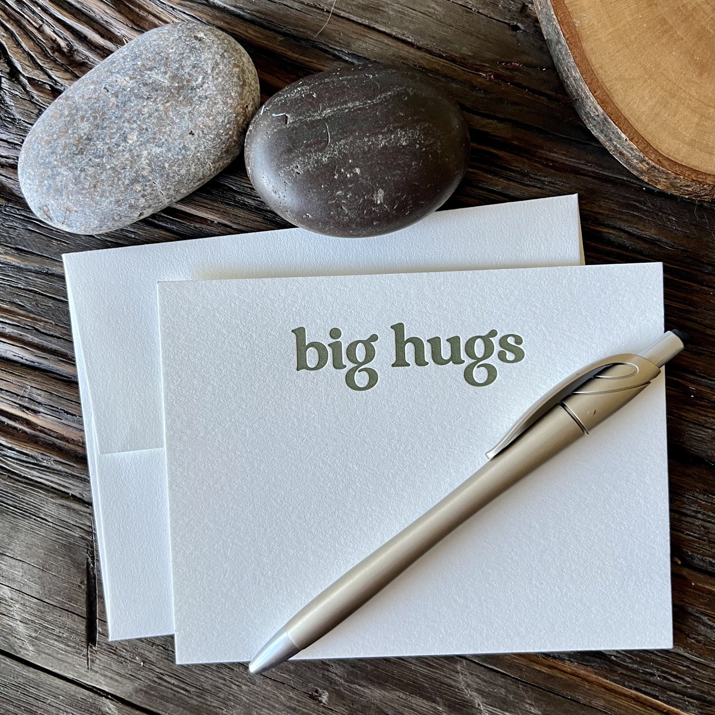 Single notecard with envelope - it says "big hugs" in olive green letterpressed ink, styled on a wooden table with river rocks, a gold pen and a sliced wood coaster.