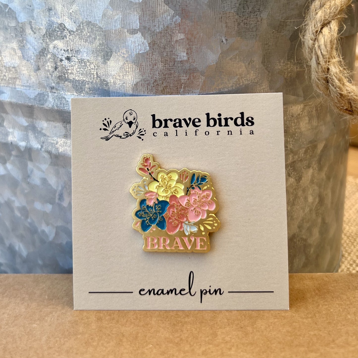 Enamel pin on a square backing card, featuring multi color flowers, gold accents and the word "BRAVE"
