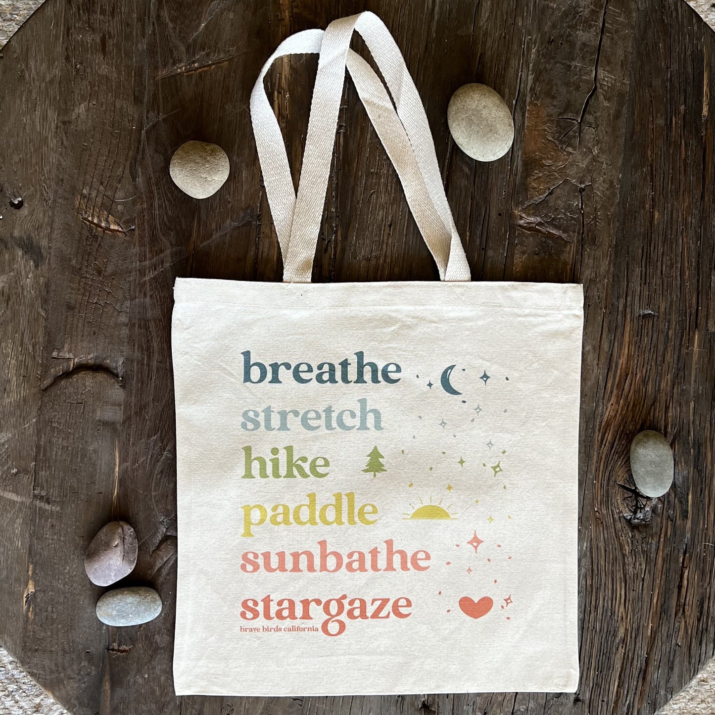 Natural Canvas Tote with rainbow colored ink and the words breathe, stretch, hike, paddle, sunbathe, stargaze, each in a different color. Shown flat on a rustic wood surface with river rocks scattered around.