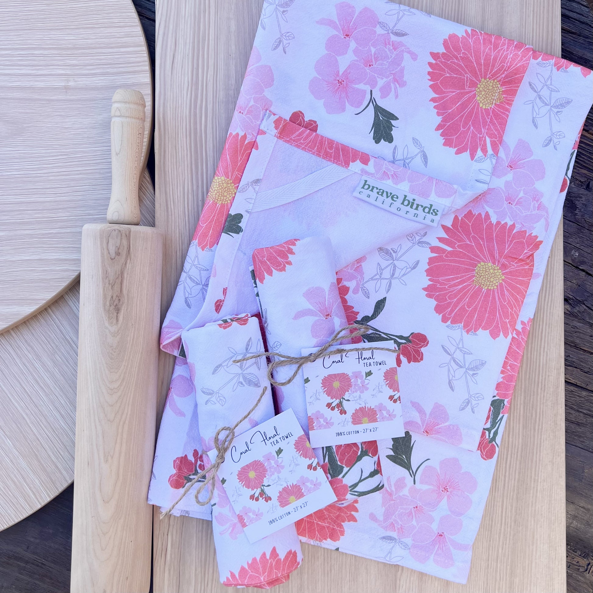 White cotton tea towels with pink floral print, one shown open with corner folded back to reveal a hanging loop and logo label, and two shown rolled up and tied with twine and a hang tag.