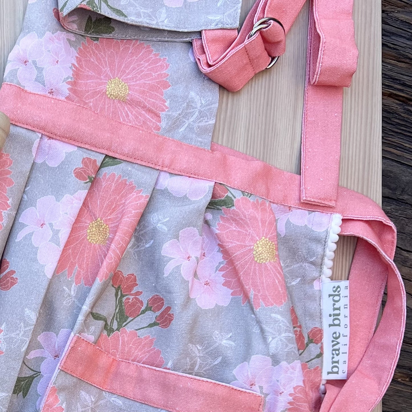 Zoomed in detail shot of apron trim: pleating at the waist, coral pink trim, woven label reading "brave birds california" and white scalloped fabric edging