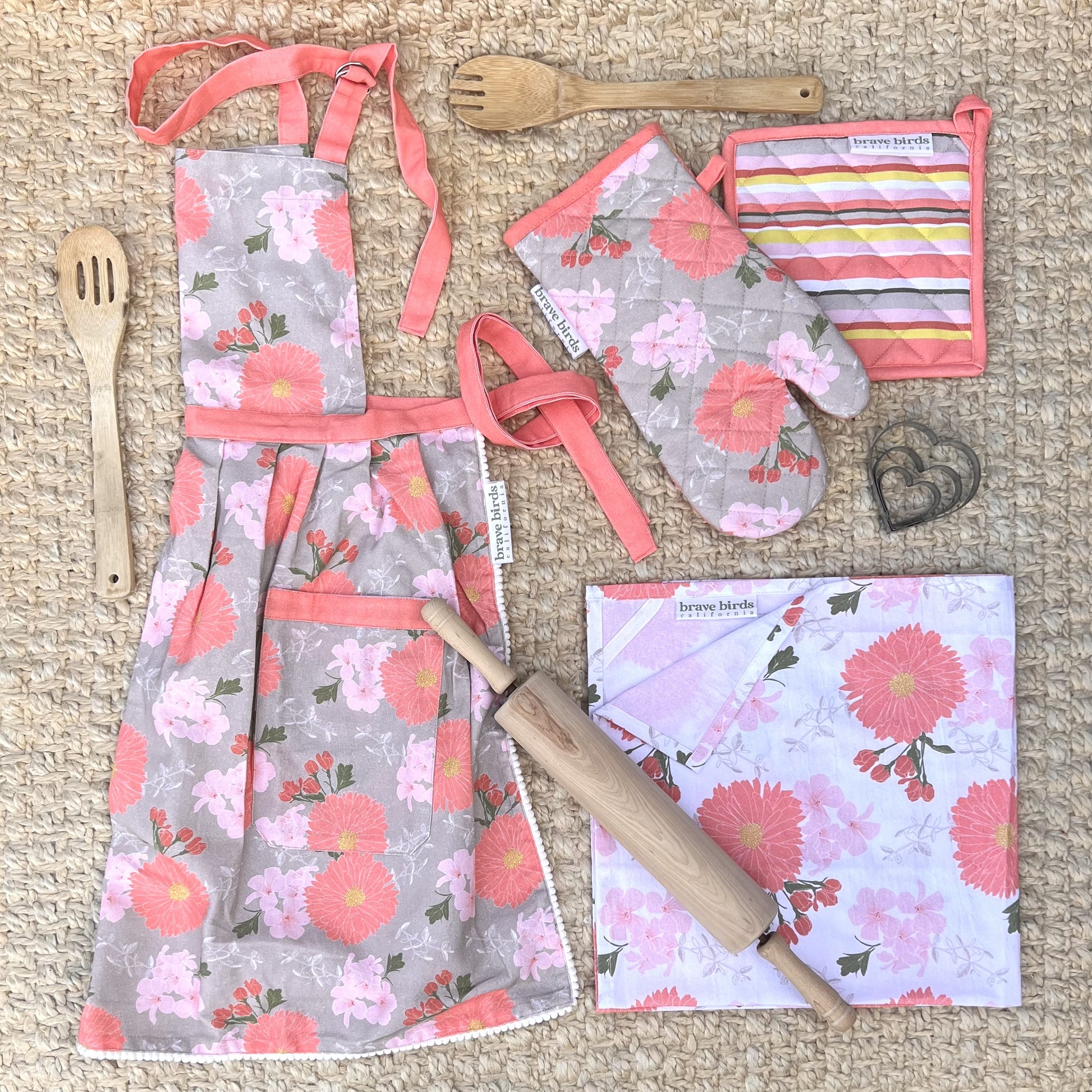 Collection of floral print kitchen textiles in pink, coral, khaki and white color palette. Apron, oven mitt, potholder and tea towel, styled with a rolling pin, wooden spoons and cookie cutters.