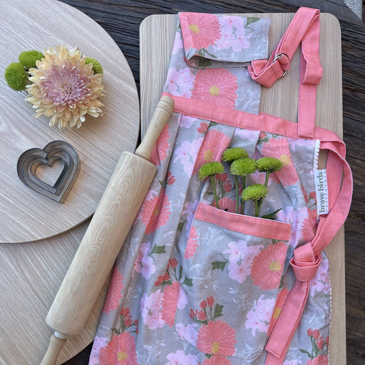 Cotton apron with a floral print in coral, pink and khaki, coral pink tie accents, styled with a rolling pin, heart shaped cookie cutters and green flowers