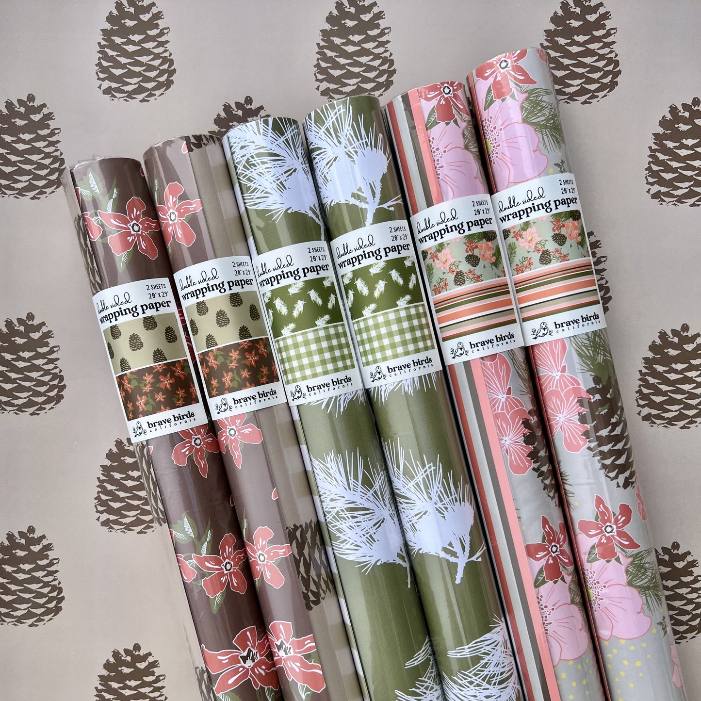 Rolls of packaged wrapping paper in plastic sleeves with sticker labels. Showing all 3 new patterns with pine, floral, stripe and gingham details