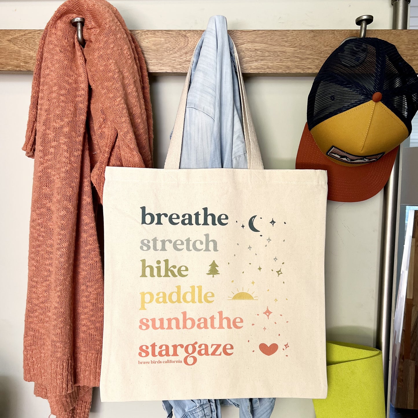 Natural Canvas Tote with rainbow colored ink and the words breathe, stretch, hike, paddle, sunbathe, stargaze, each in a different color. Shown hanging with a sweater and a hat on metal hooks in an entryway coat rack.