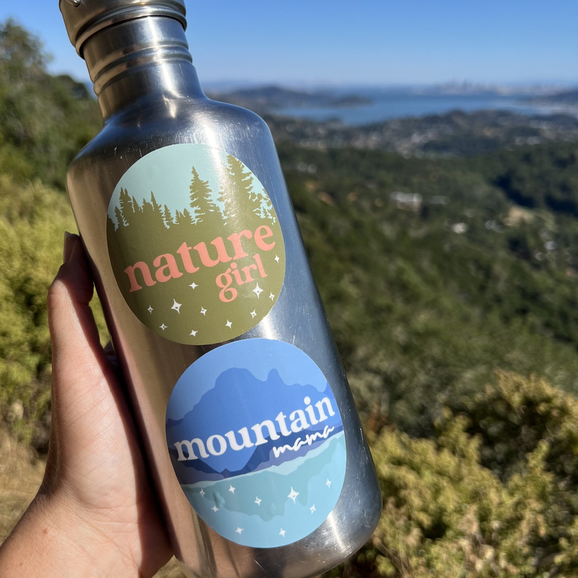 hand holding a water bottle with two circular stickers on it, in front of an outdoor scene with mountains and water in the background