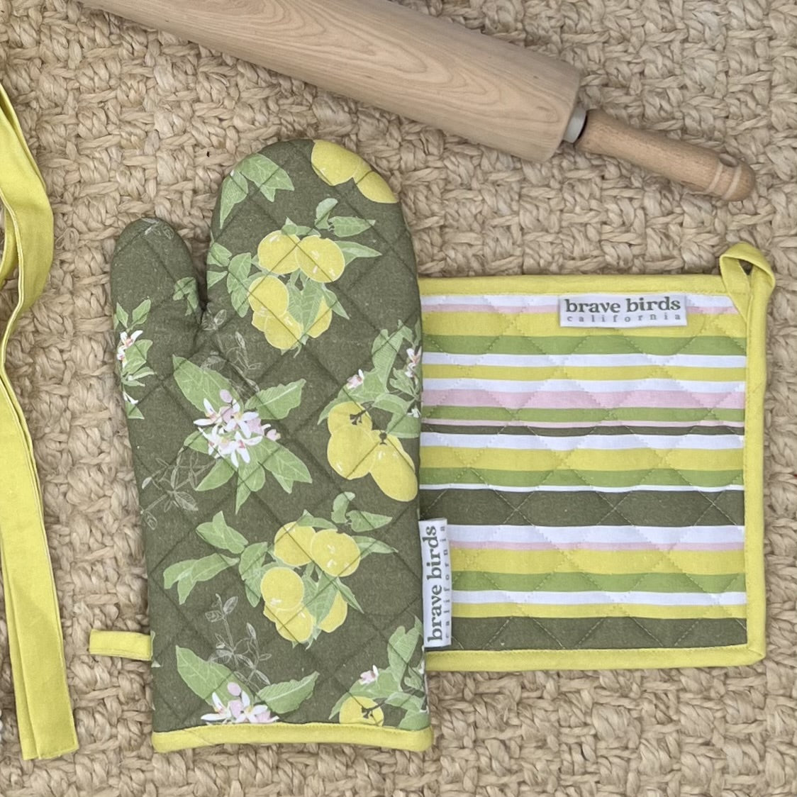Lemon and floral printed oven mitt in olive green & yellow with a color-coordinated striped square potholder, both with hanging loops, styled with a wooden rolling pin