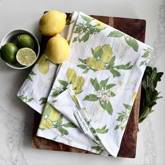 Lemon print tea towel on a wood cutting board, styled with fresh lemons, limes and mint