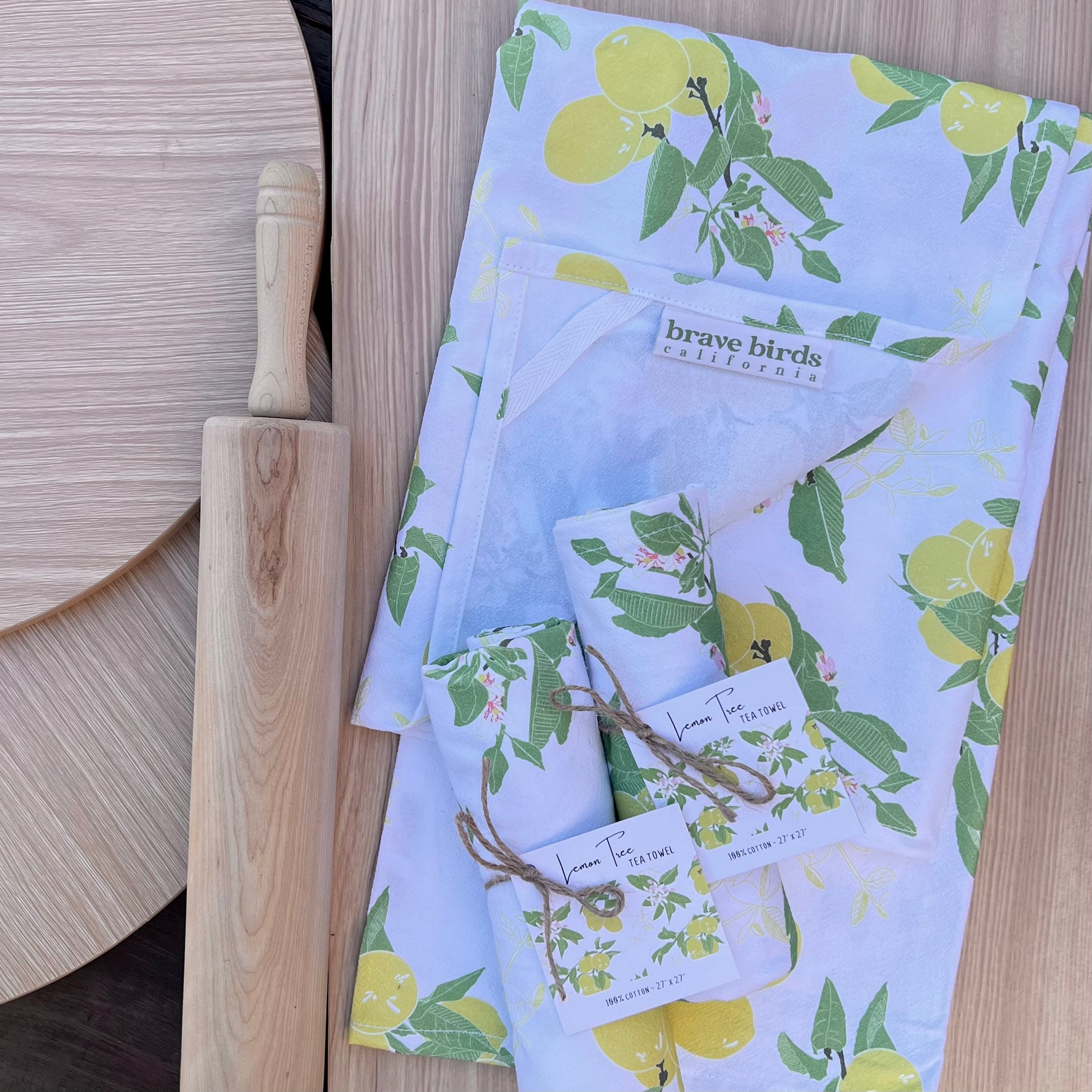 White cotton tea towels with green and yellow lemon tree print, one shown open with corner folded back to reveal a hanging loop and logo label, and two shown rolled up and tied with twine and a hang tag.