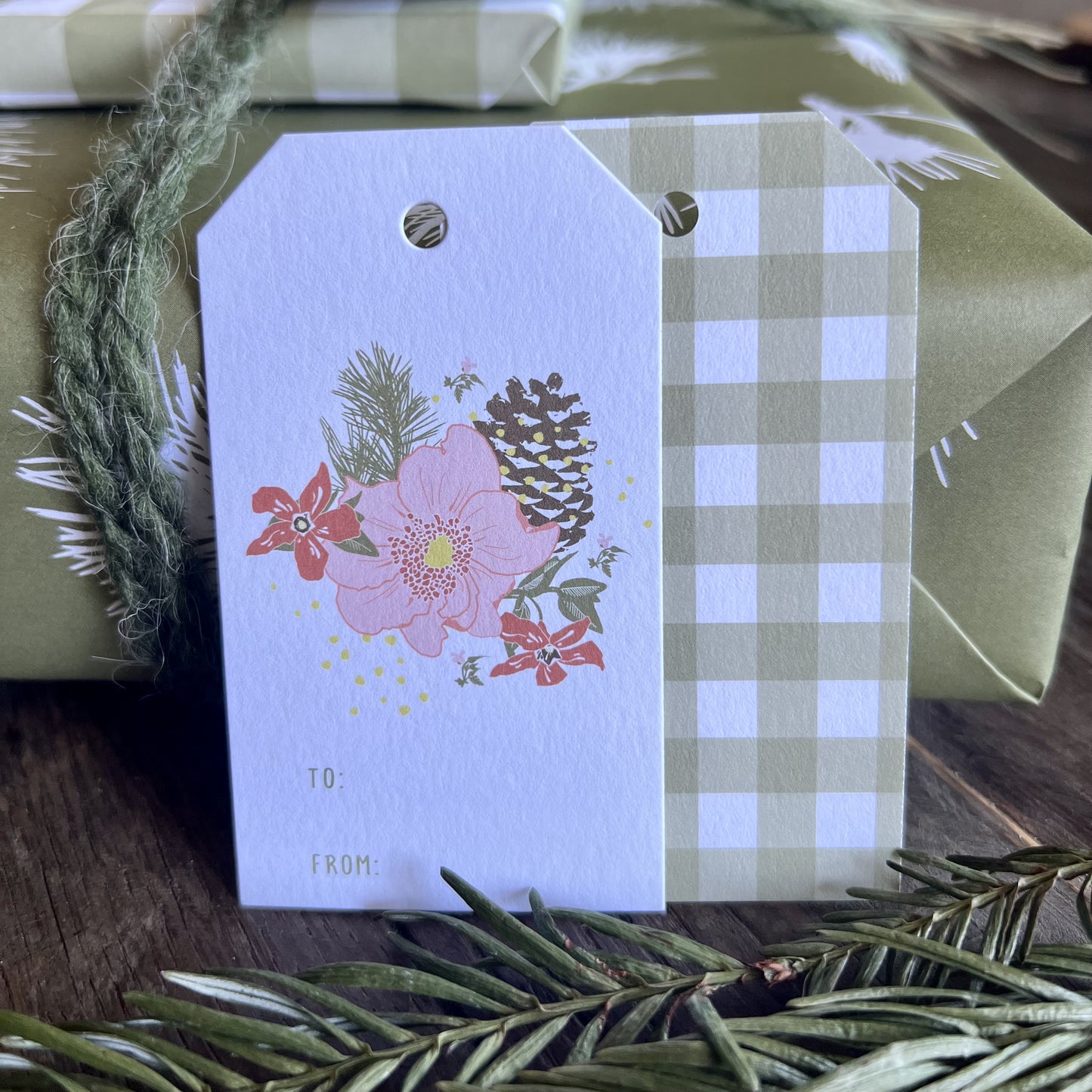 Closeup shot of a gift wrapped package and two TO/FROM gift tags with a floral and pinecone illustration on the front and a green gingham plaid print on the back..