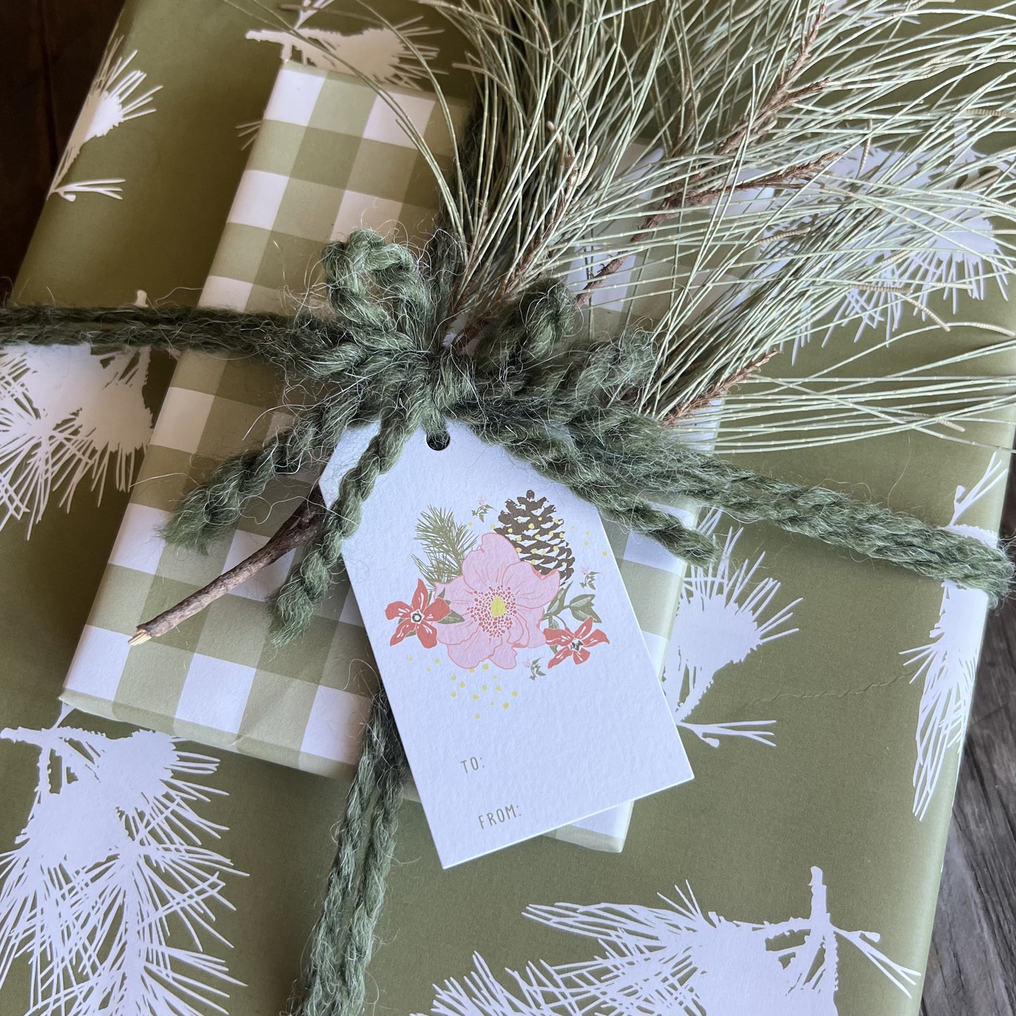 Gift wrapped package tied with green yarn and a TO/FROM gift tag with a floral and pinecone illustration.