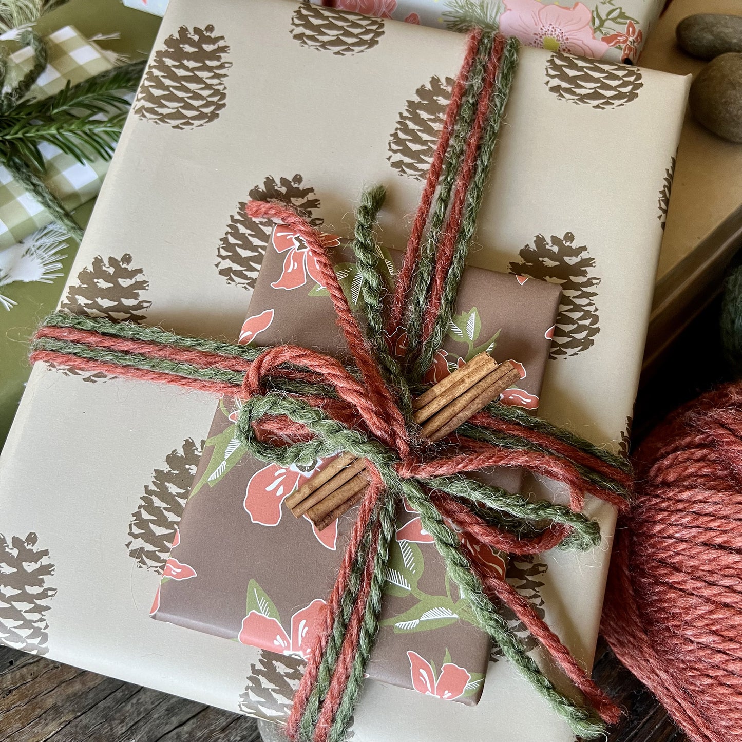 Giftwrapped package with two boxes, one in a khaki and brown pinecone pattern, one in a red and brown floral print, tied with red and green yarn