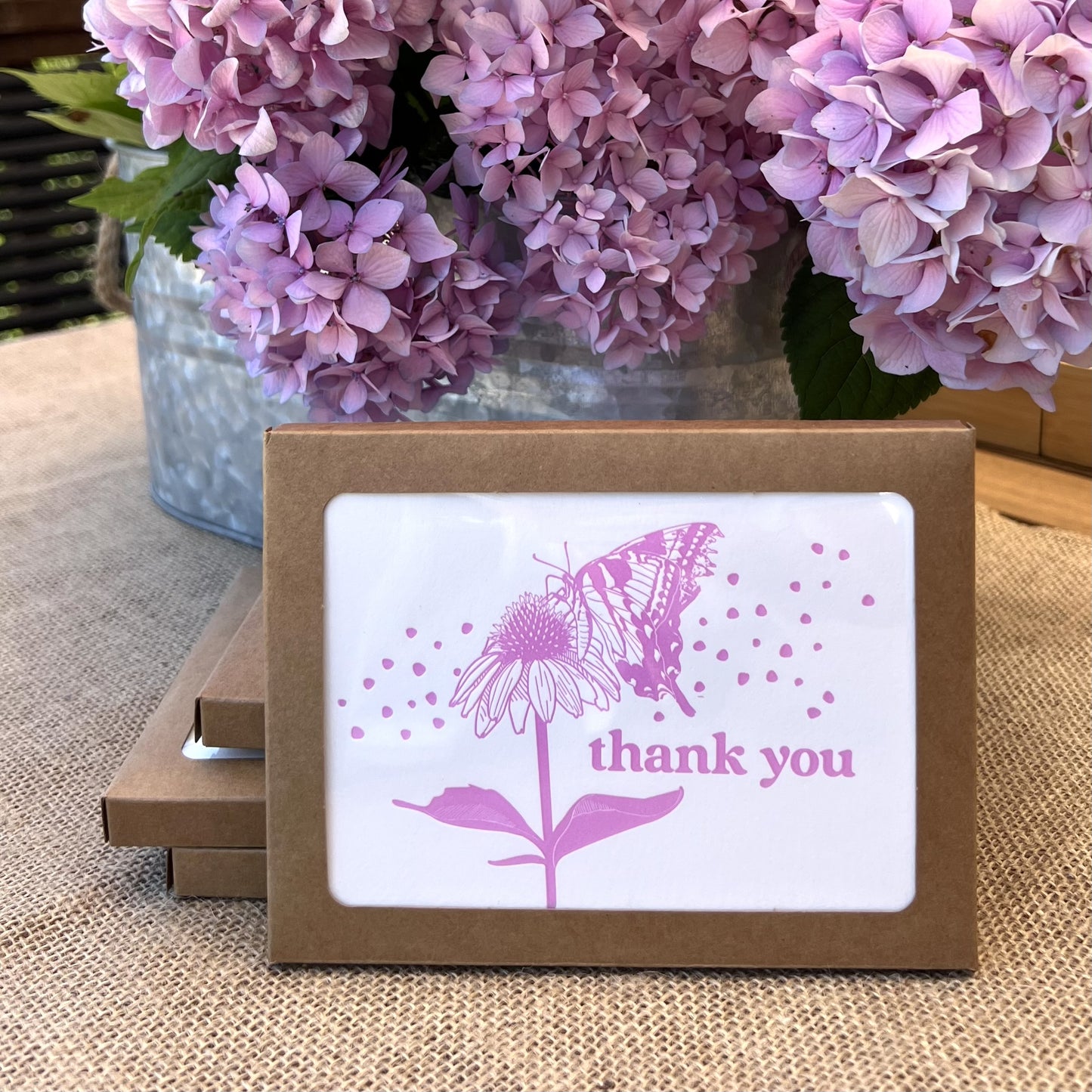 Stack of kraft paper boxes with a window showing a dark pink print of a butterfly, echinacea flower and the words "thank you" styled in front of a bouquet of hydrangeas in matching dark pink