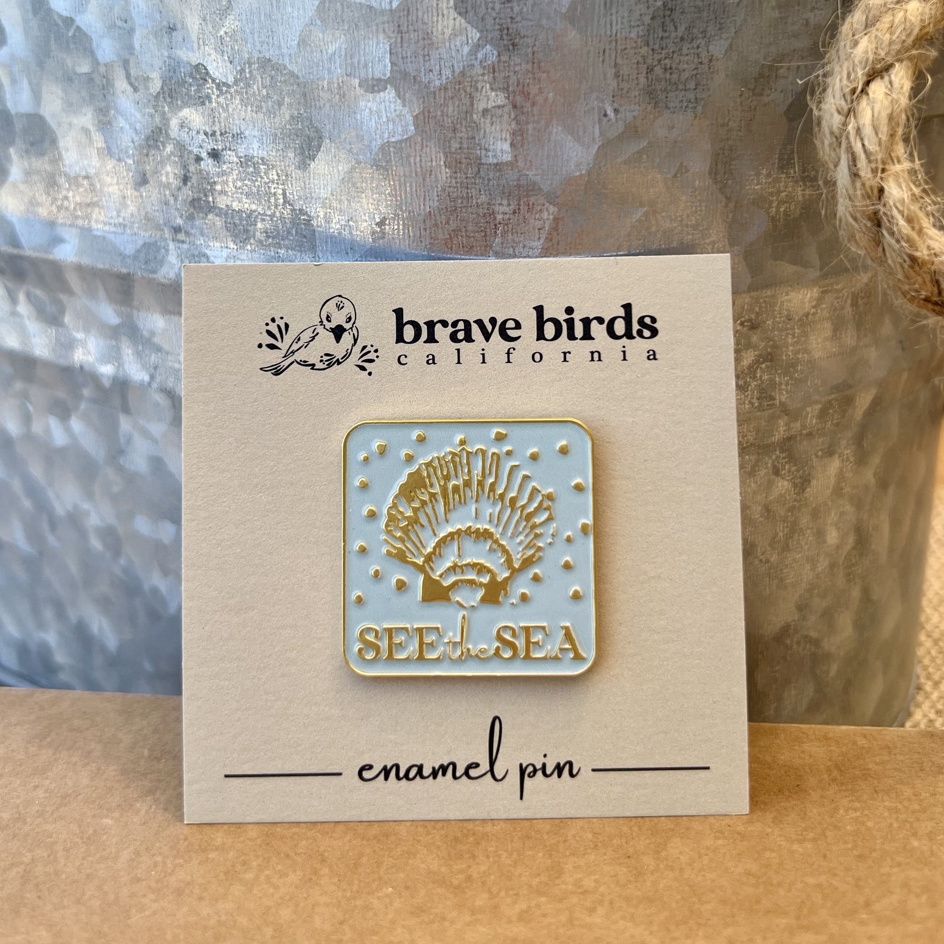 Square, light blue enamel pin with gold metal accents. Features a scallop shell design and the words "see the sea" with a tan backing card