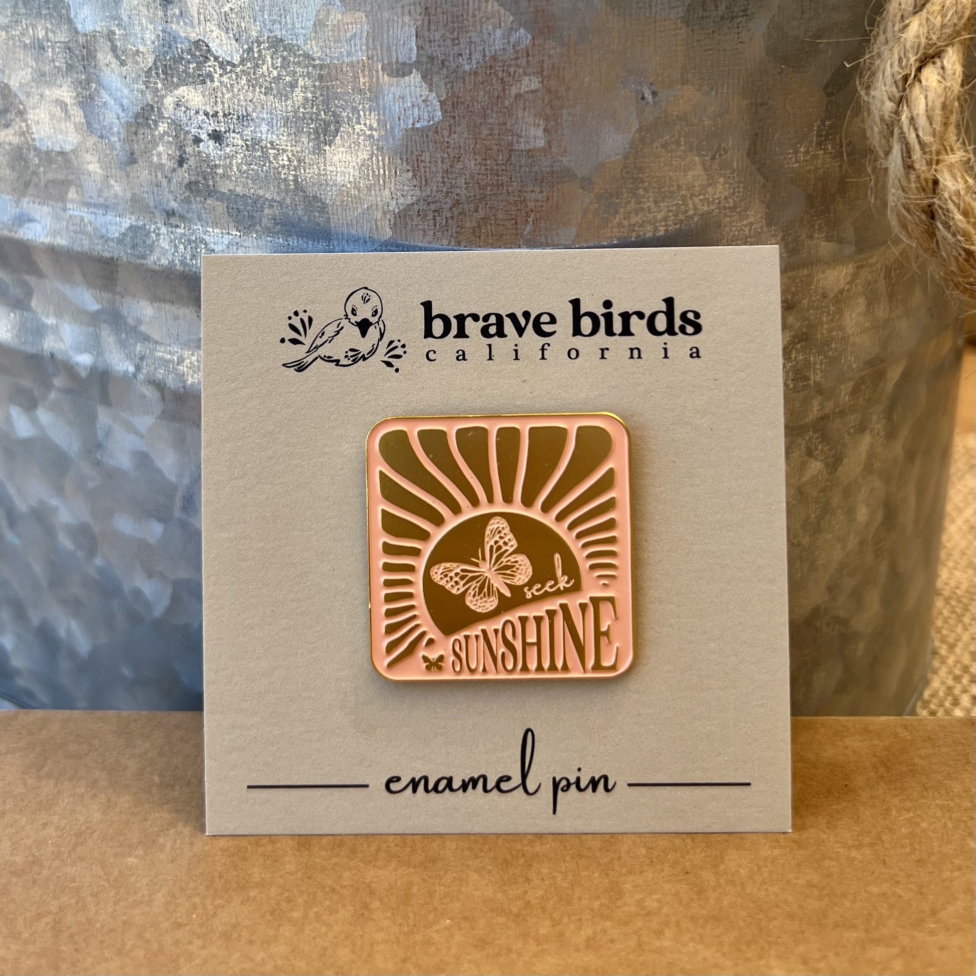 Square enamel pin with light pink and gold design featuring sun rays, a butterfly and the words "seek sunshine" shown on a square branded backing card