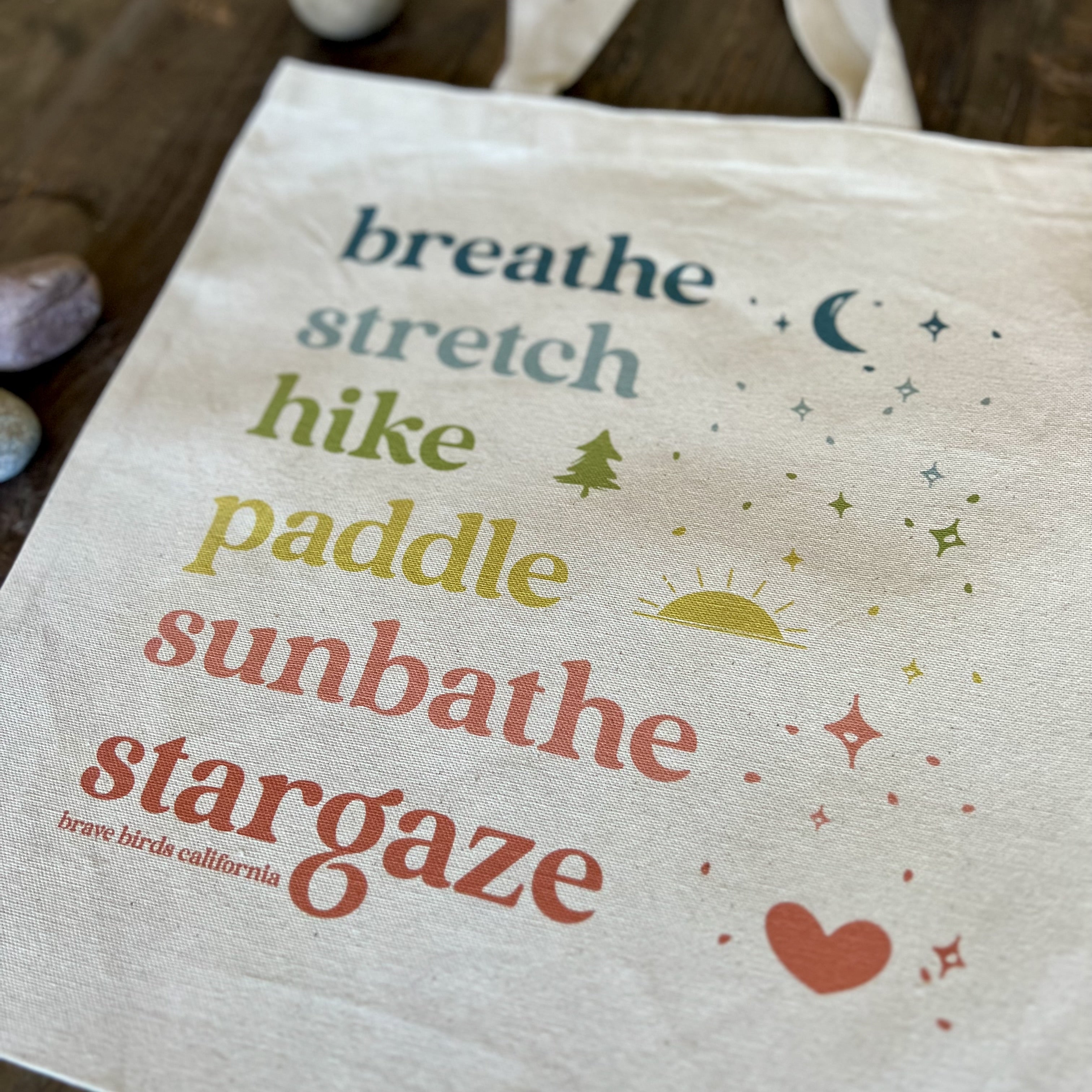 Closeup detail shot of the print on a natural canvas tote with rainbow colored ink and the words breathe, stretch, hike, paddle, sunbathe, stargaze, each in a different color.
