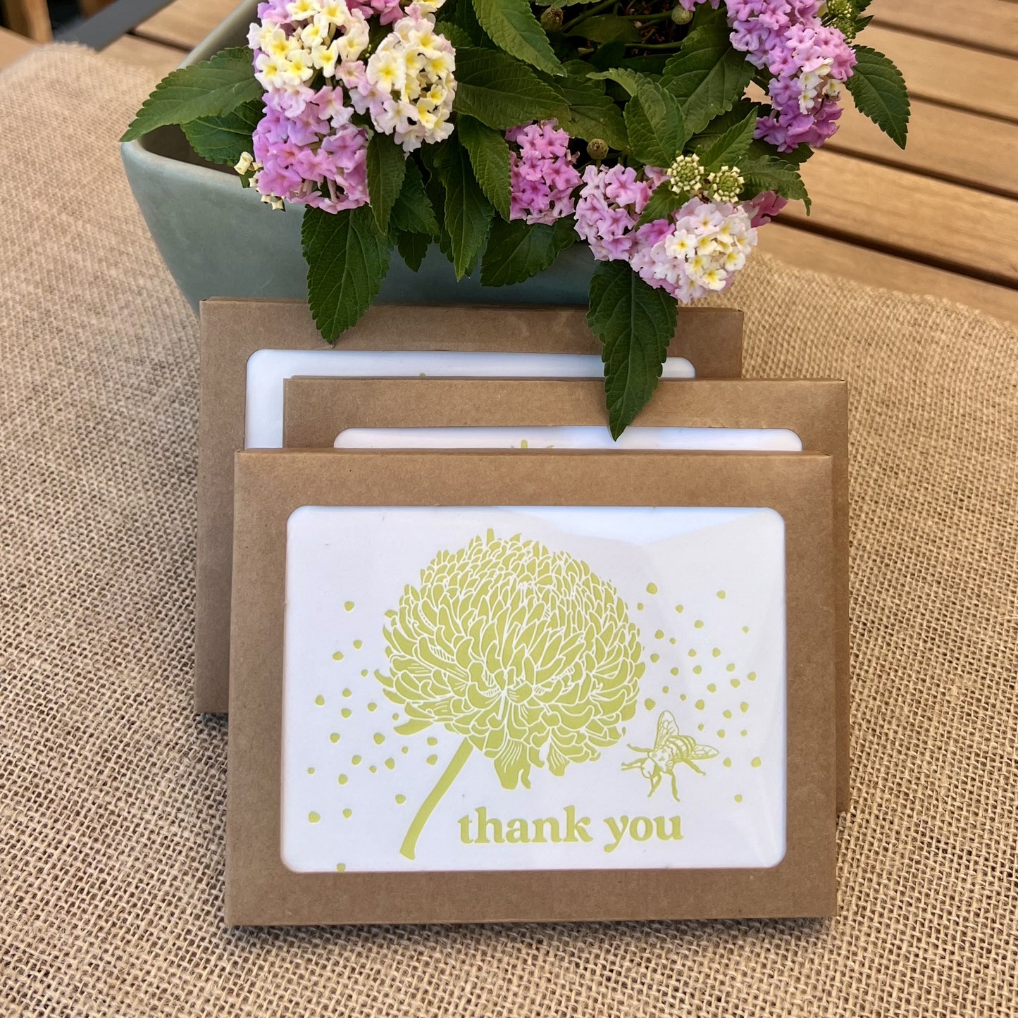 Stack of kraft paper boxes with windows showing cards with citron yellow flowers and bees, with text reading "thank you", styled with fresh pink hydrangea flowers