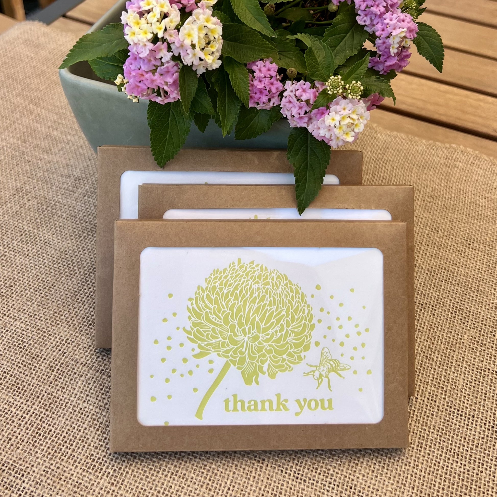 Stack of kraft paper boxes with windows showing cards with citron yellow flowers and bees, with text reading "thank you", styled with fresh pink hydrangea flowers