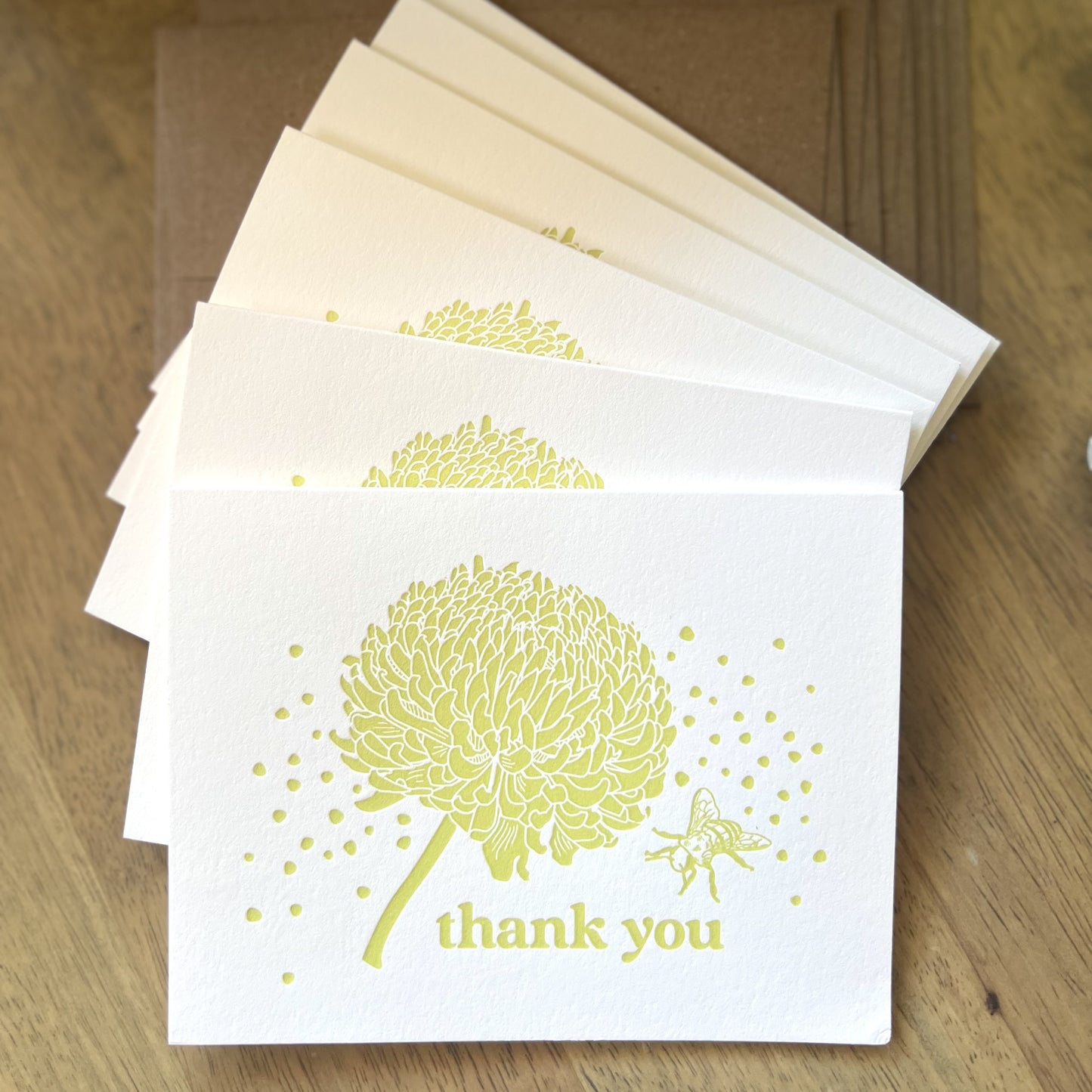 Stack of six letterpress cards with citron yellow ink, featuring an illustration of a chrysanthemum flowere and a bee and the words "thank you", shown with a stack of kraft paper brown envelopes