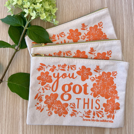 Stack of three zip pouches in natural canvas with bright orange print and gold zippers. Print reads "you got this"