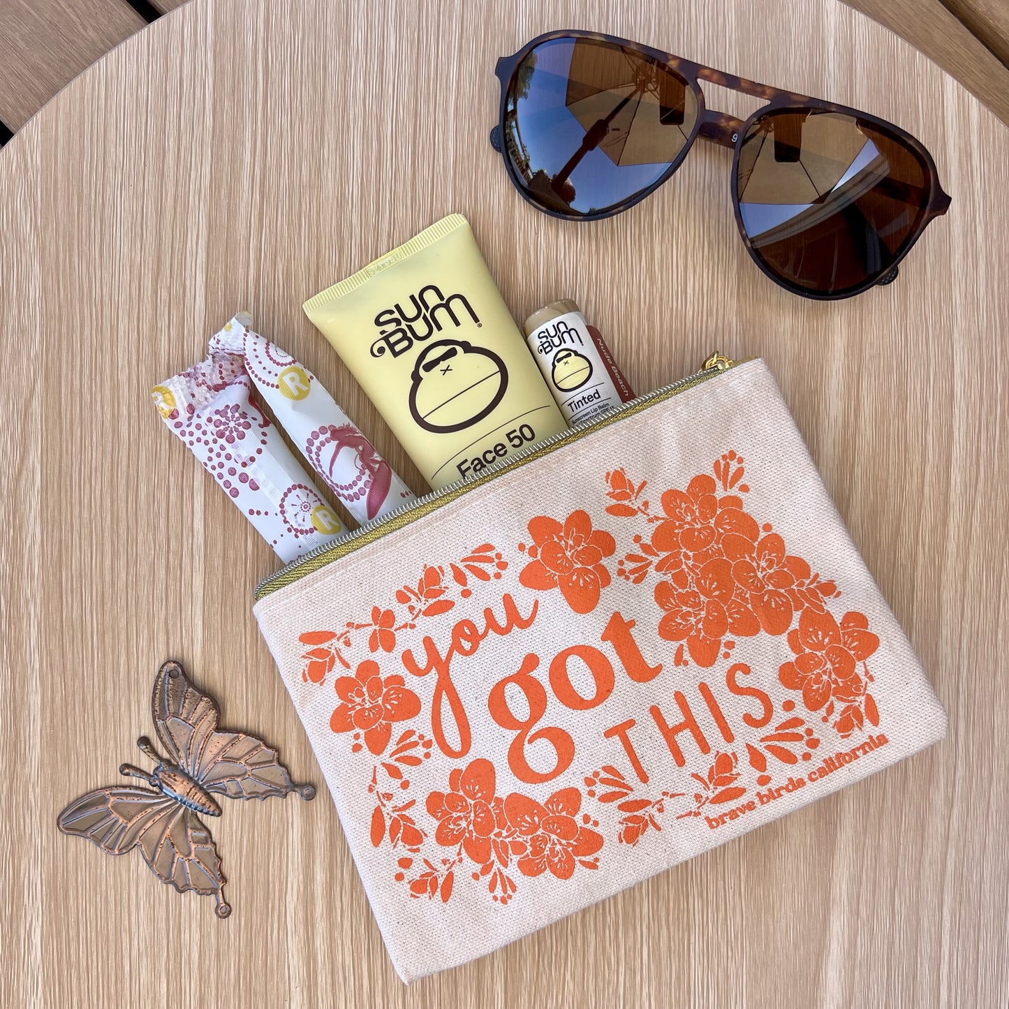 Zip pouch in natural canvas with bright orange print and gold zippers, shown open with summer accessories peeking out. Print reads "you got this"