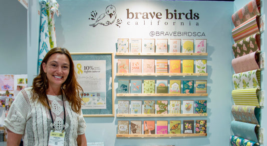 Photo of a woman wiht brown hair standing in front of a display wall with greeting cards and wrapping paper, and a logo for brave birds california