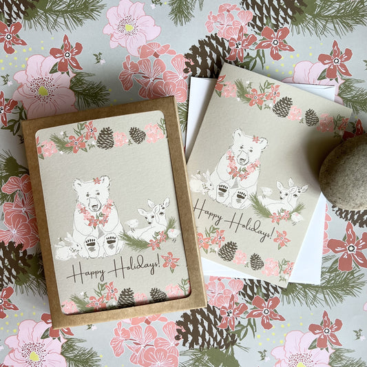 Holiday cards with a kraft paper brown background and illustrations of a bear, deer, bunny, birds and a snail, all decorated with festive flowers. Text reads "Happy Holidays" and photo shows both a boxed set and a single card with white envelope.