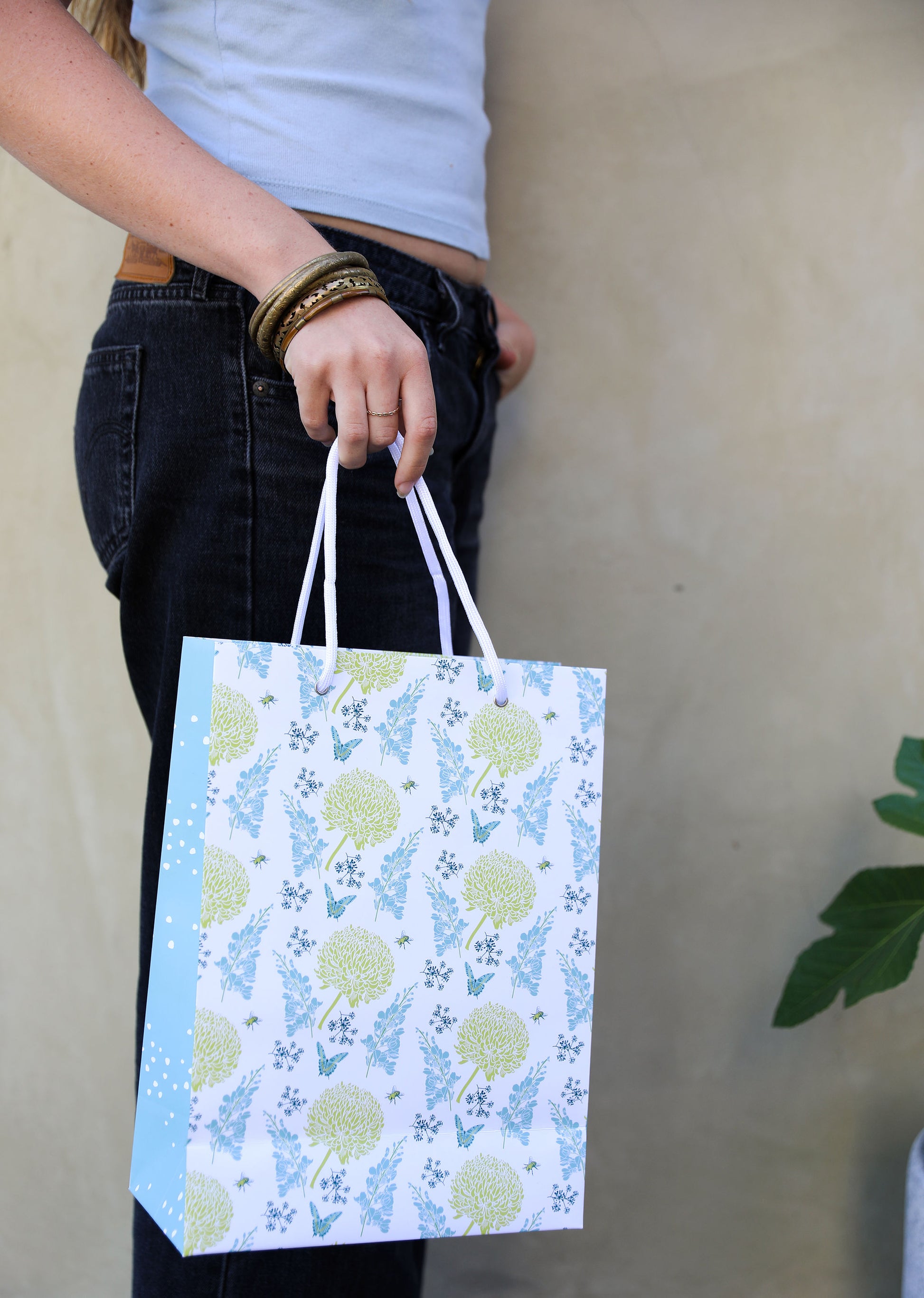 A woman in jeans holding a patterned gift bag with blue and yellow floral design on the front and a subtle blue dot on the side panel