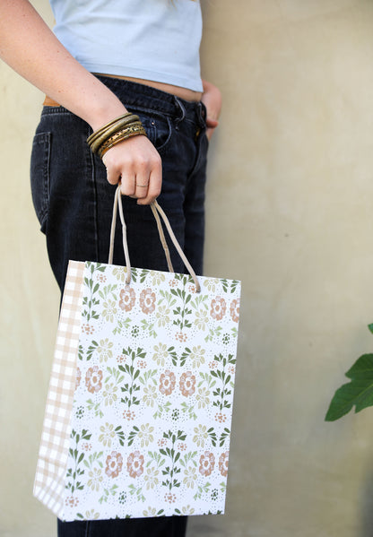 Woman in jeans holding a patterned gift bag with a tan and green floral print on the front and a tan gingham plaid pattern on the side panel