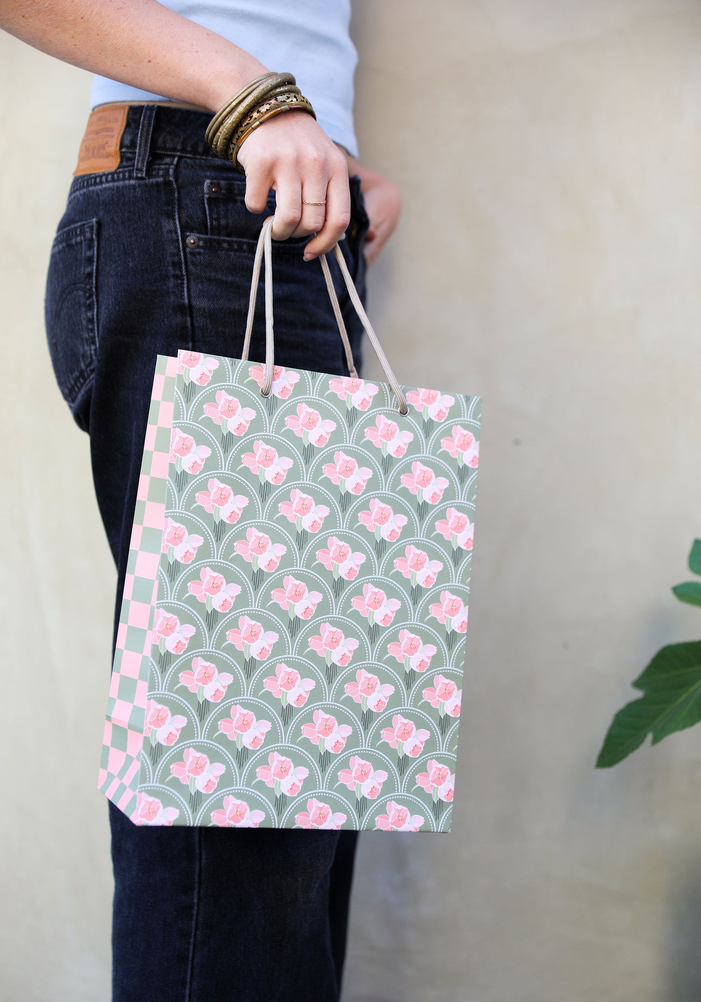 Woman in jeans holding a patterned gift bag with a pink floral scallop design on the front and a pink and green check design on the side panel