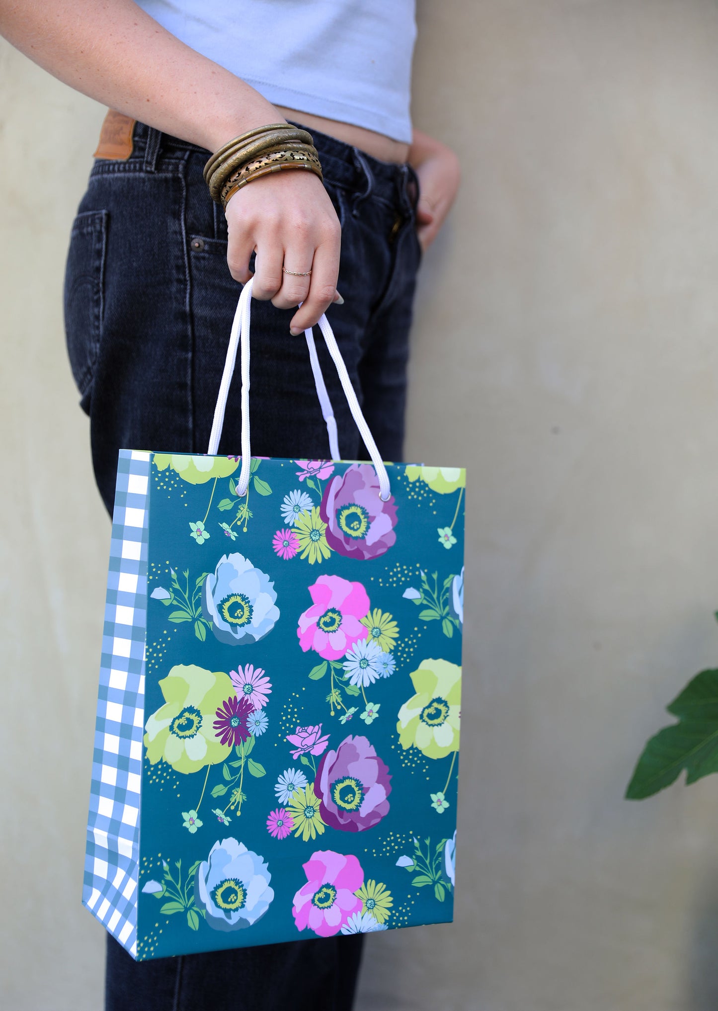 Woman in jeans holding a patterned gift bag with a teal grounded floral print on the front and a teal gingham plaid on the side panel 